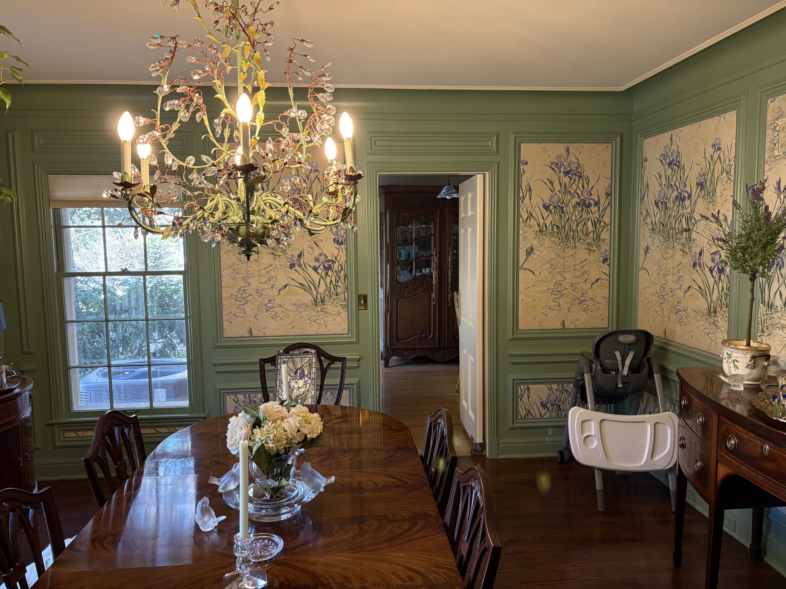 A dining room with green walls and floral wallpaper, a large chandelier with crystal accents hanging above a wooden dining table, which has a floral centerpiece and candles. There is a window with blinds, a wooden china cabinet, and various furniture