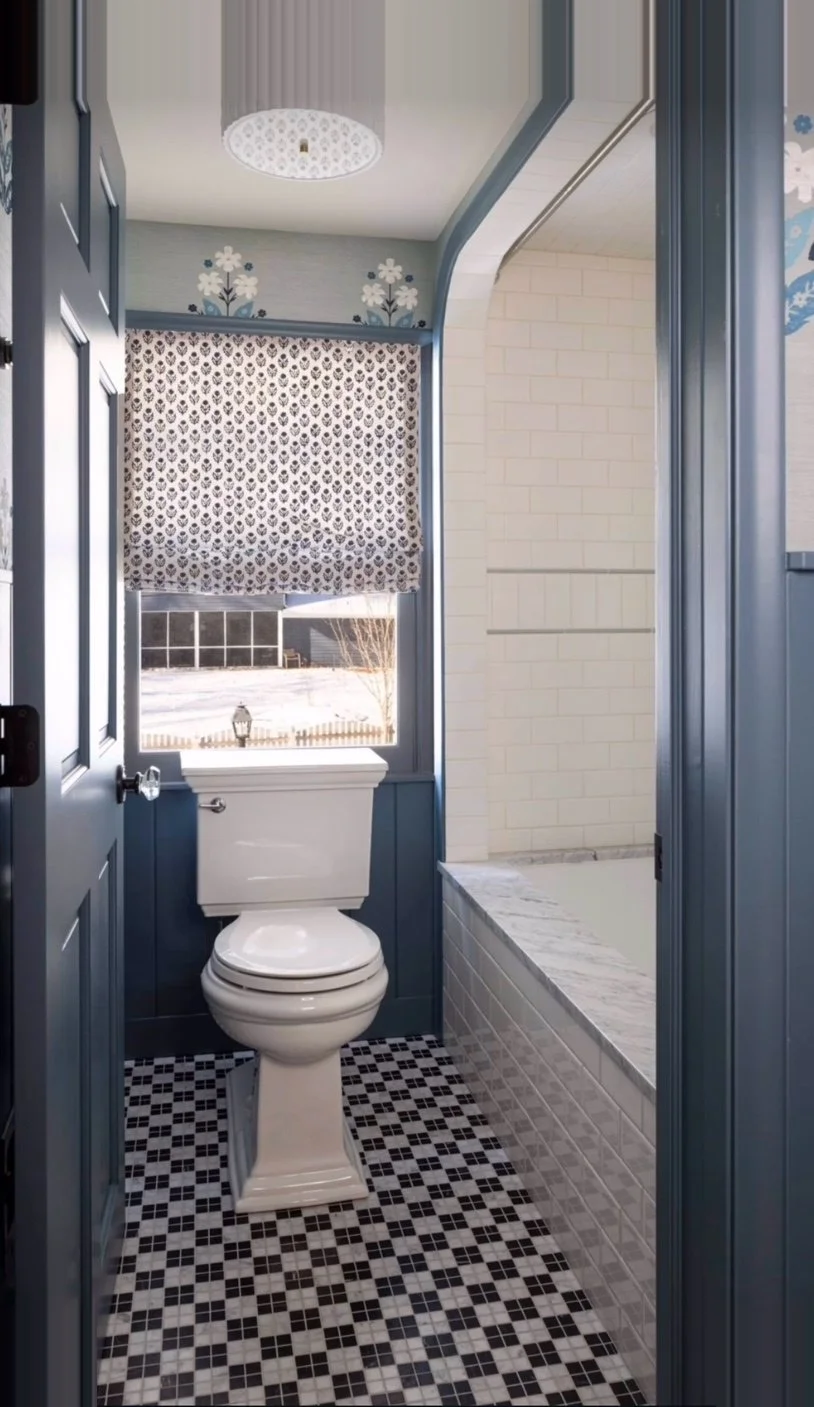 View of a bathroom with a toilet beneath a window; the window has a patterned curtain, and there is a bathtub with a marble ledge to the right. The bathroom has blue walls, a white ceiling, and a black and white checkered floor.
