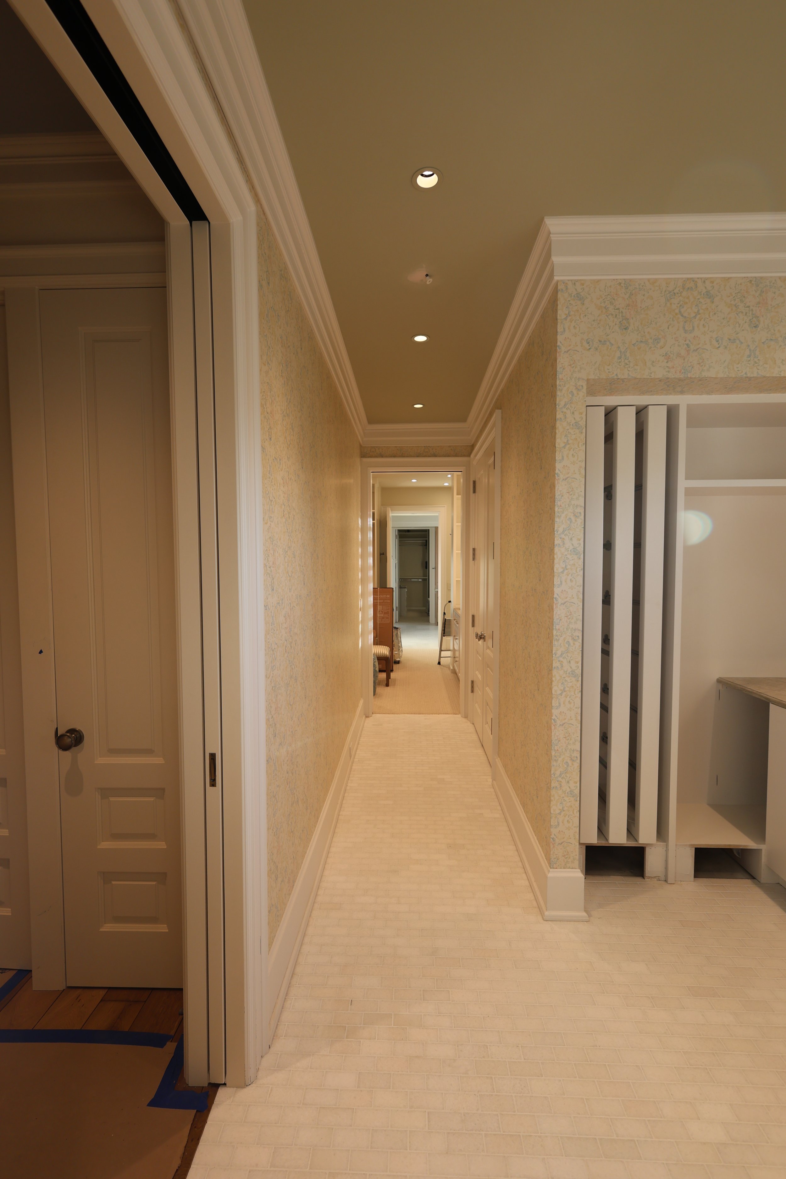 A hallway in a house with cream-colored walls, white crown molding, and a beige tiled floor. The hallway extends into another room in the background, with part of a chair visible on the left side.