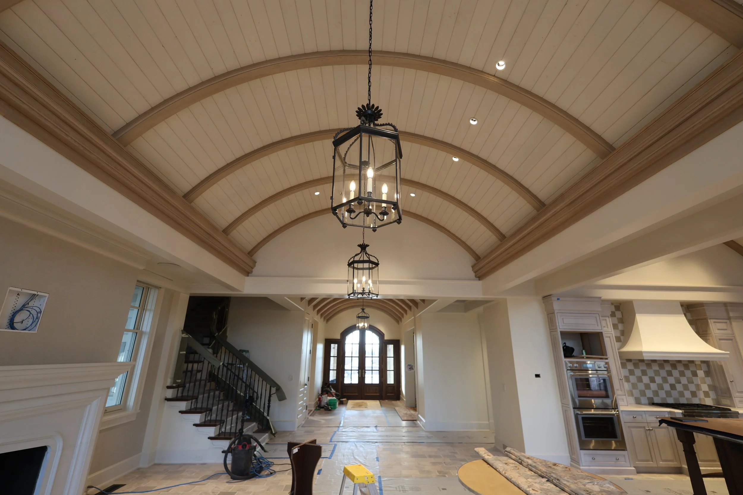 Interior view of a house under construction, showing a high ceiling with arched wooden panels, hanging lantern-style chandeliers, and a staircase on the left side leading up to the second floor.