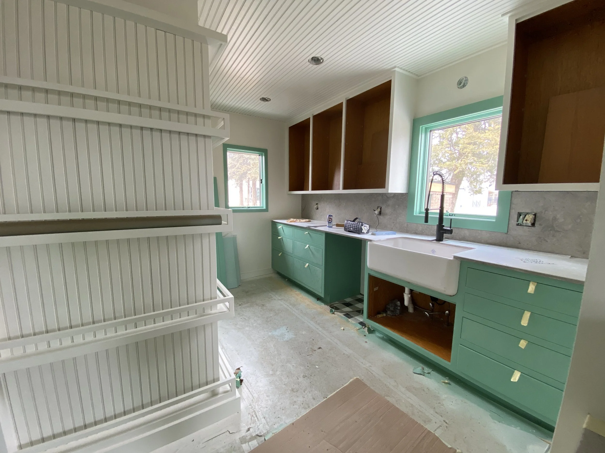 Interior of a kitchen under construction with green lower cabinets, open upper cabinets, a white farmhouse sink, large windows, and unfinished flooring.