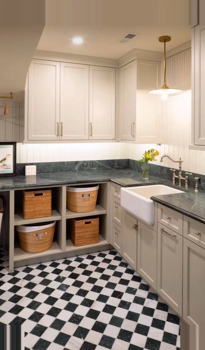 Kitchen with white cabinets, black and white checkered floor, farmhouse sink, black marble countertops, and woven baskets in open shelving.