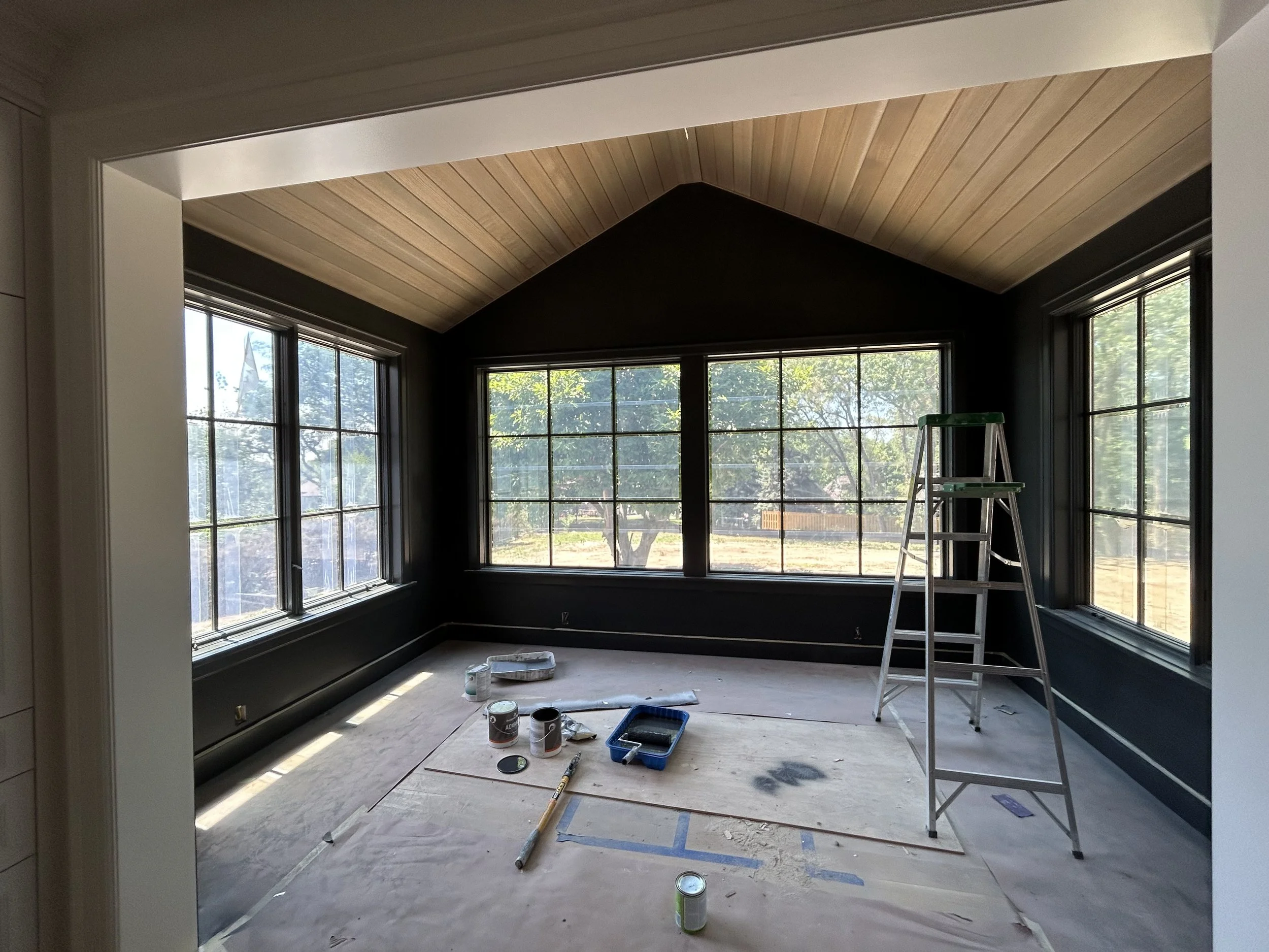 Room under renovation with large windows, ladder, and paint supplies on the floor.