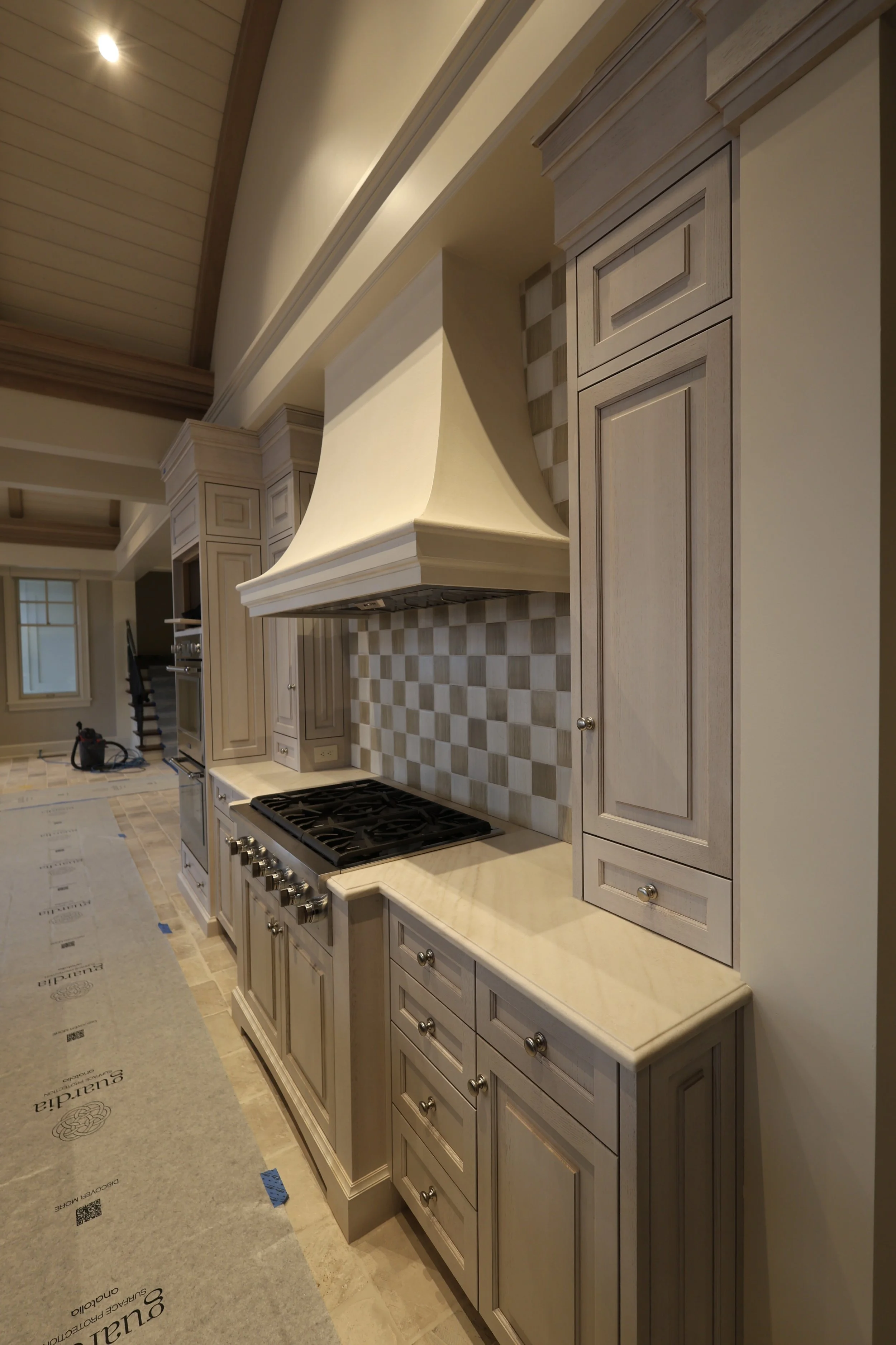 Kitchen with beige cabinets, a beige countertop, a stove with knobs, a tiled backsplash with checkered pattern, and a range hood overhead. There is a staircase visible in the background and kitchen flooring with protective covering.