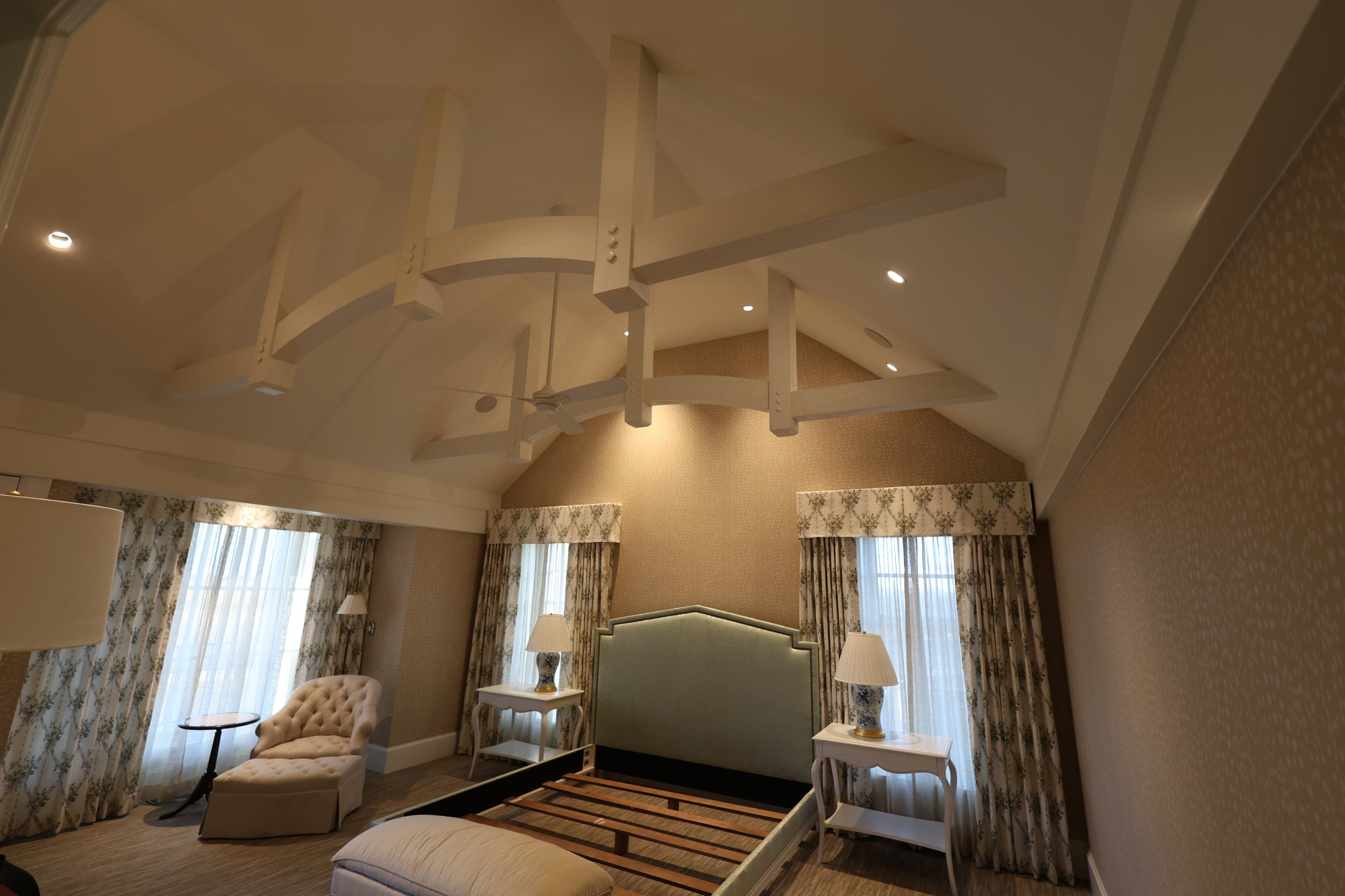 A luxurious hotel room with a high vaulted ceiling featuring white wooden beams and a ceiling fan. The room has large windows with floral curtains, a tufted armchair with a small round table, a bed with a tufted headboard, and two matching nightstand