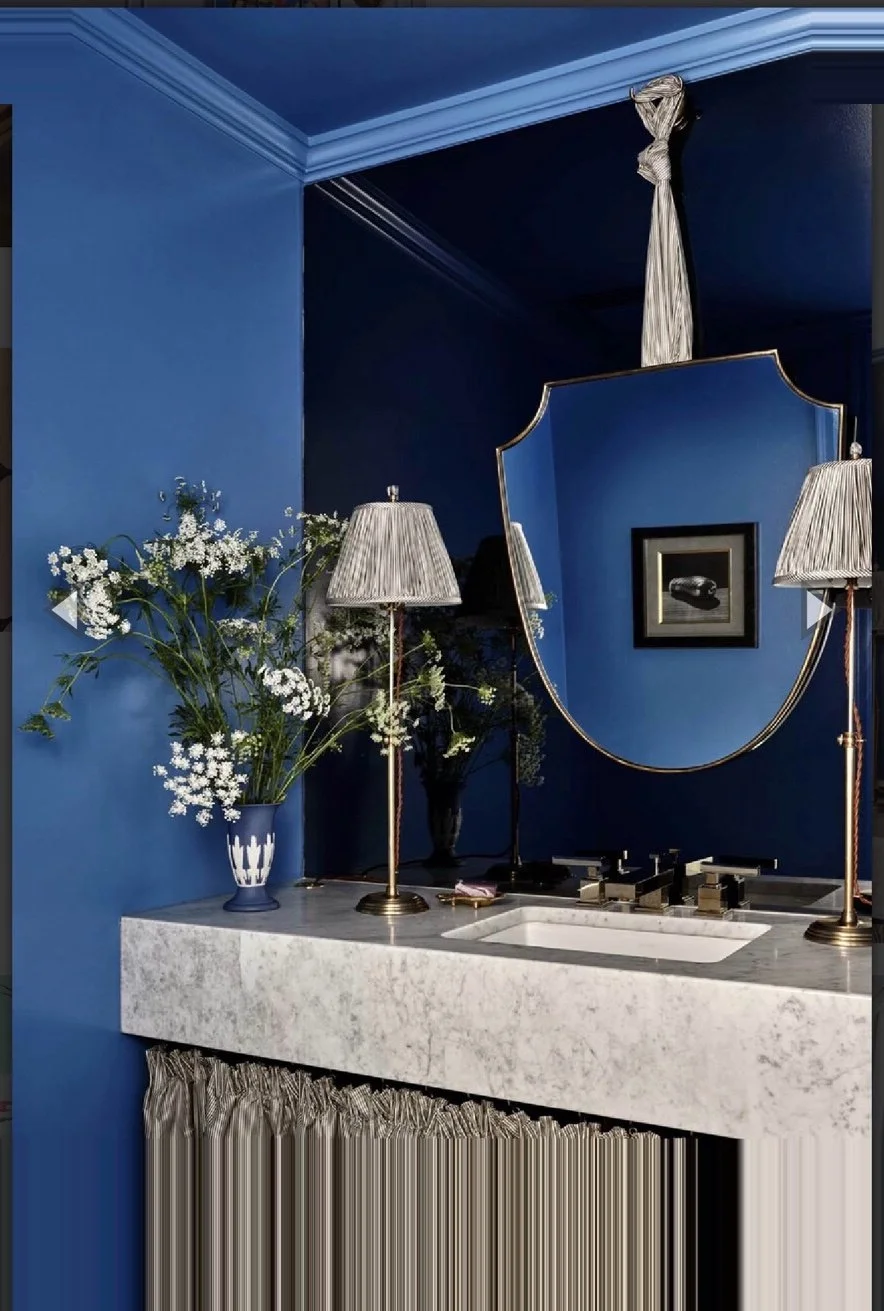 A bathroom vanity with a marble countertop, a vase of white flowers, a large decorative mirror, and two tall lamps with pleated shades, all set against a deep blue wall.