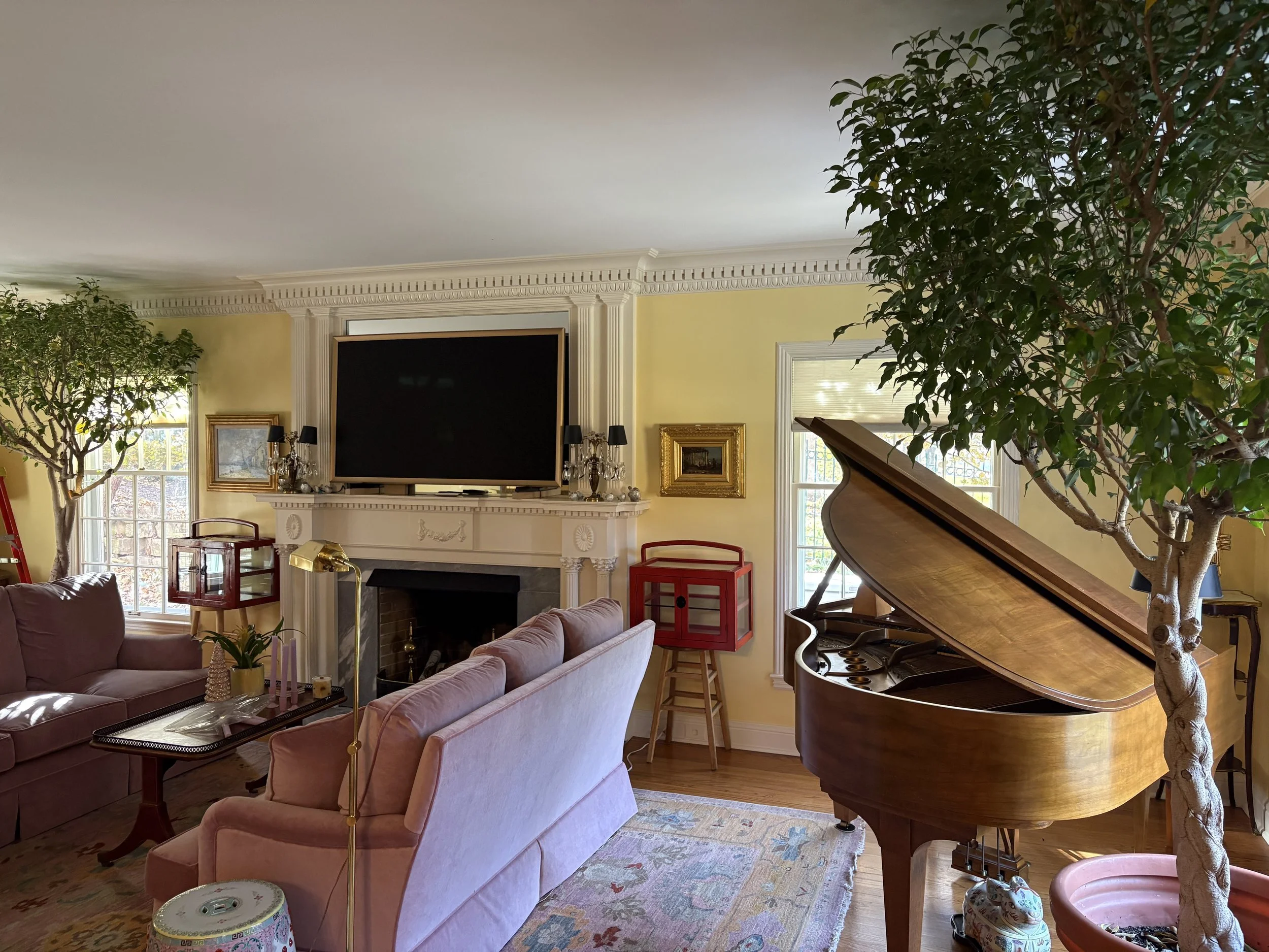Interior of a living room with a pink sofa, a grand piano, two large trees inside, a fireplace with a TV above, and various decorations including framed artwork and lamps.