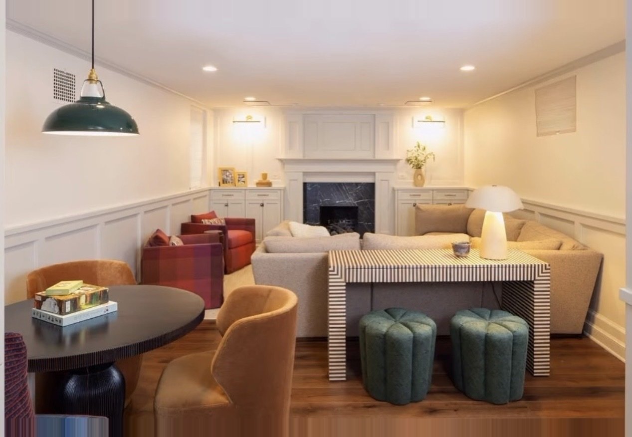 A cozy living room with a fireplace, beige sofas, a black round dining table with upholstered chairs, and decorative items including a lamp, vases, and framed photos.