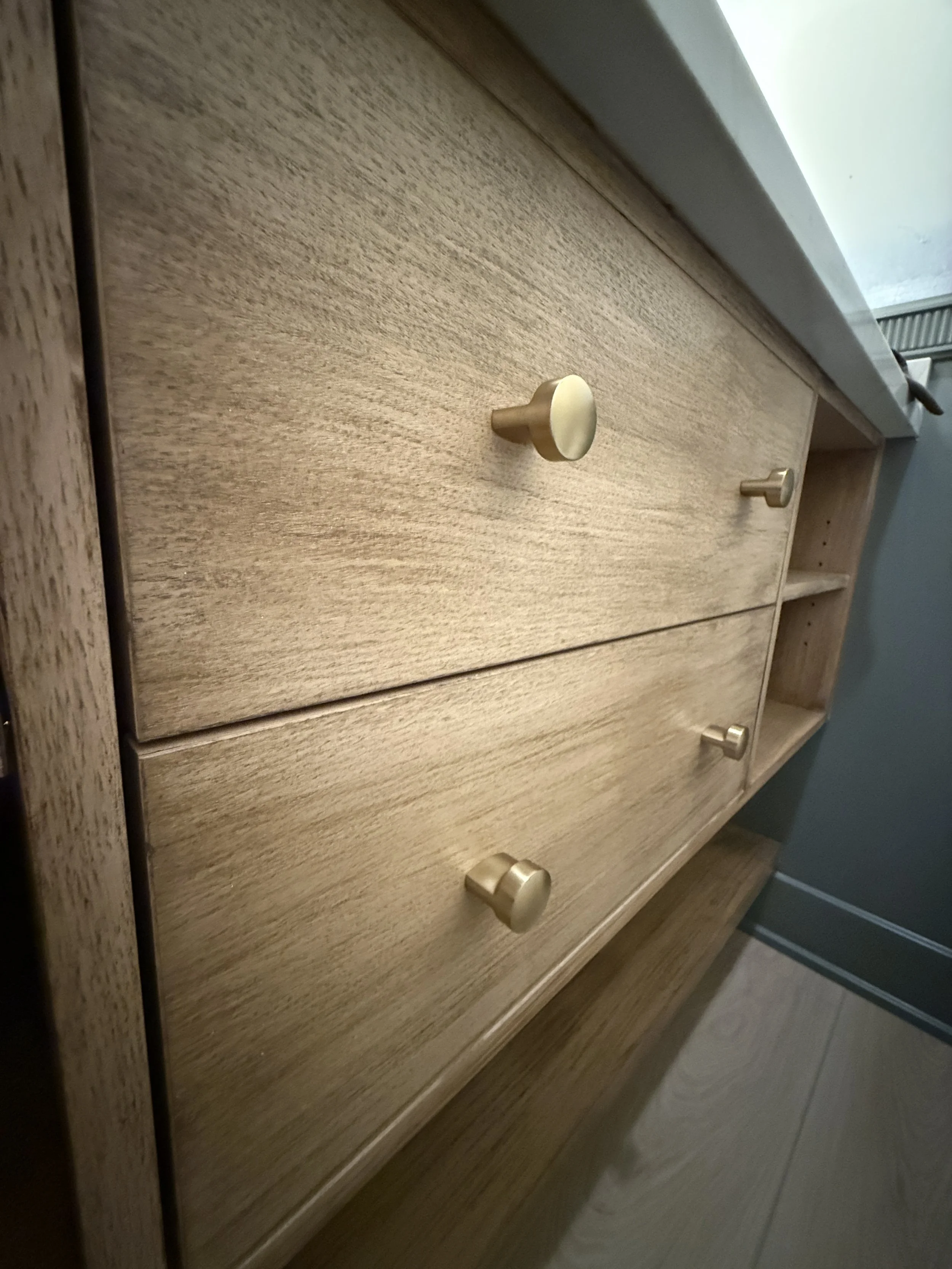 Close-up of a light-colored wooden drawer with round metallic knobs, part of a wooden dresser, next to a white countertop and a vent in the background.