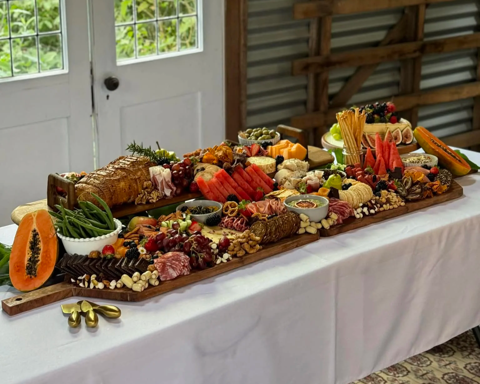 A large cheese and charcuterie board on a white tablecloth, featuring sliced meats, various cheeses, fresh fruits like watermelon, papaya, grapes, and berries, nuts, crackers, and olives, with a background of a rustic wooden interior and white door with glass panes.