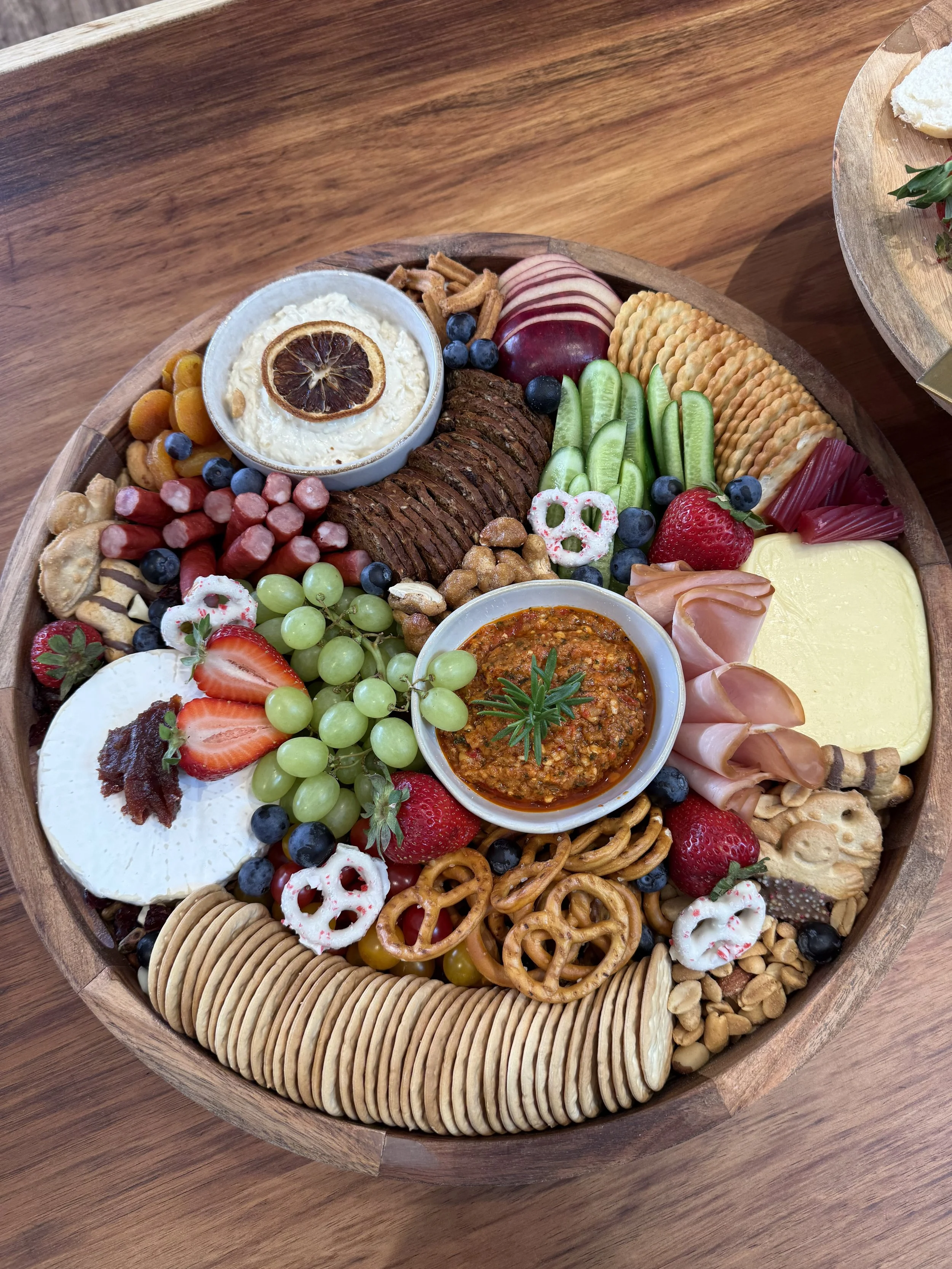 A large wooden platter with assorted snacks, including crackers, fruits like strawberries, grapes, blueberries, and sliced apple, cheese, meat slices, dips, pretzels, cookies, and nuts.