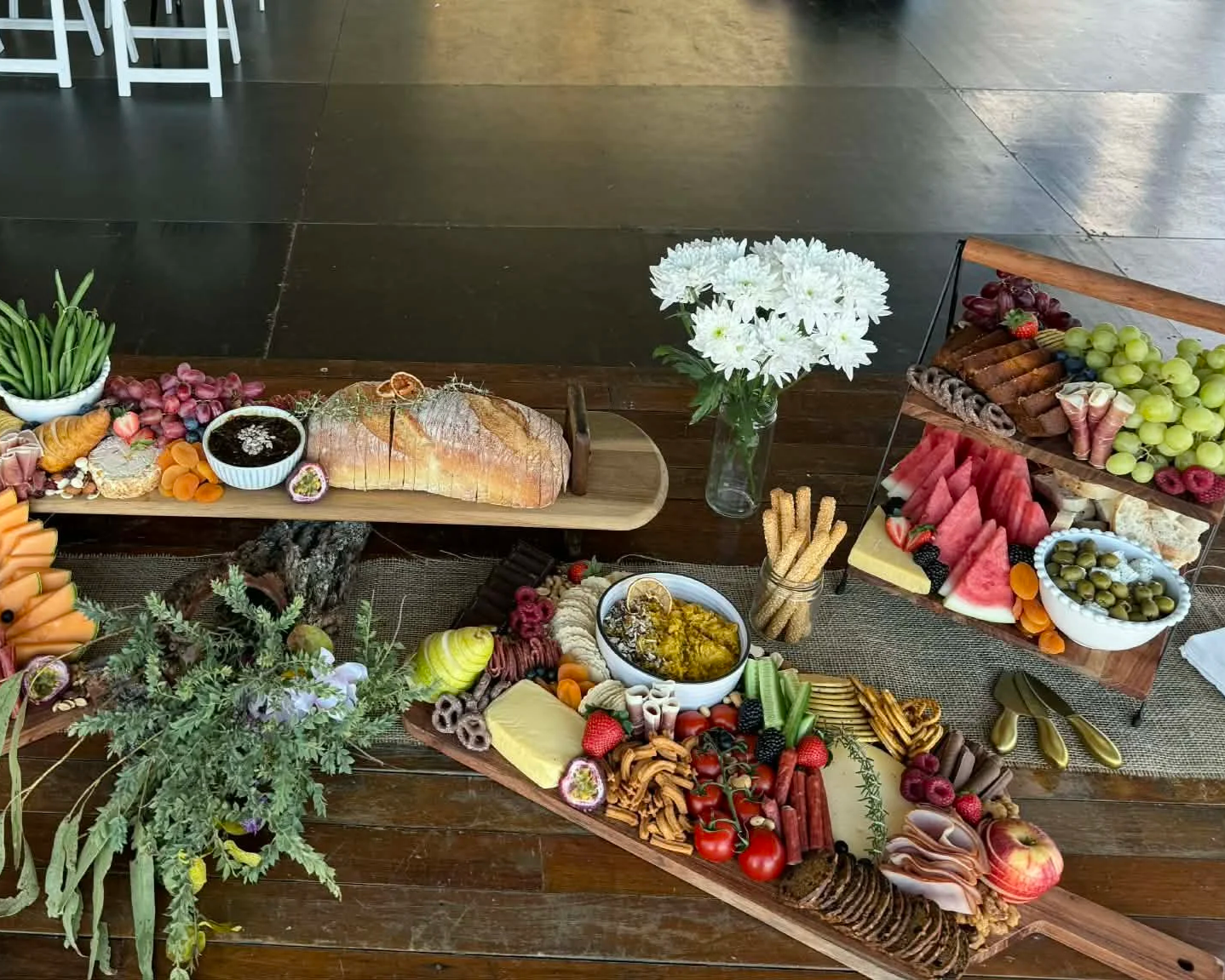 A spread of assorted cheeses, fruits, vegetables, baked goods, and snacks on a wooden table and tiered tray, with a vase of white flowers in the center.