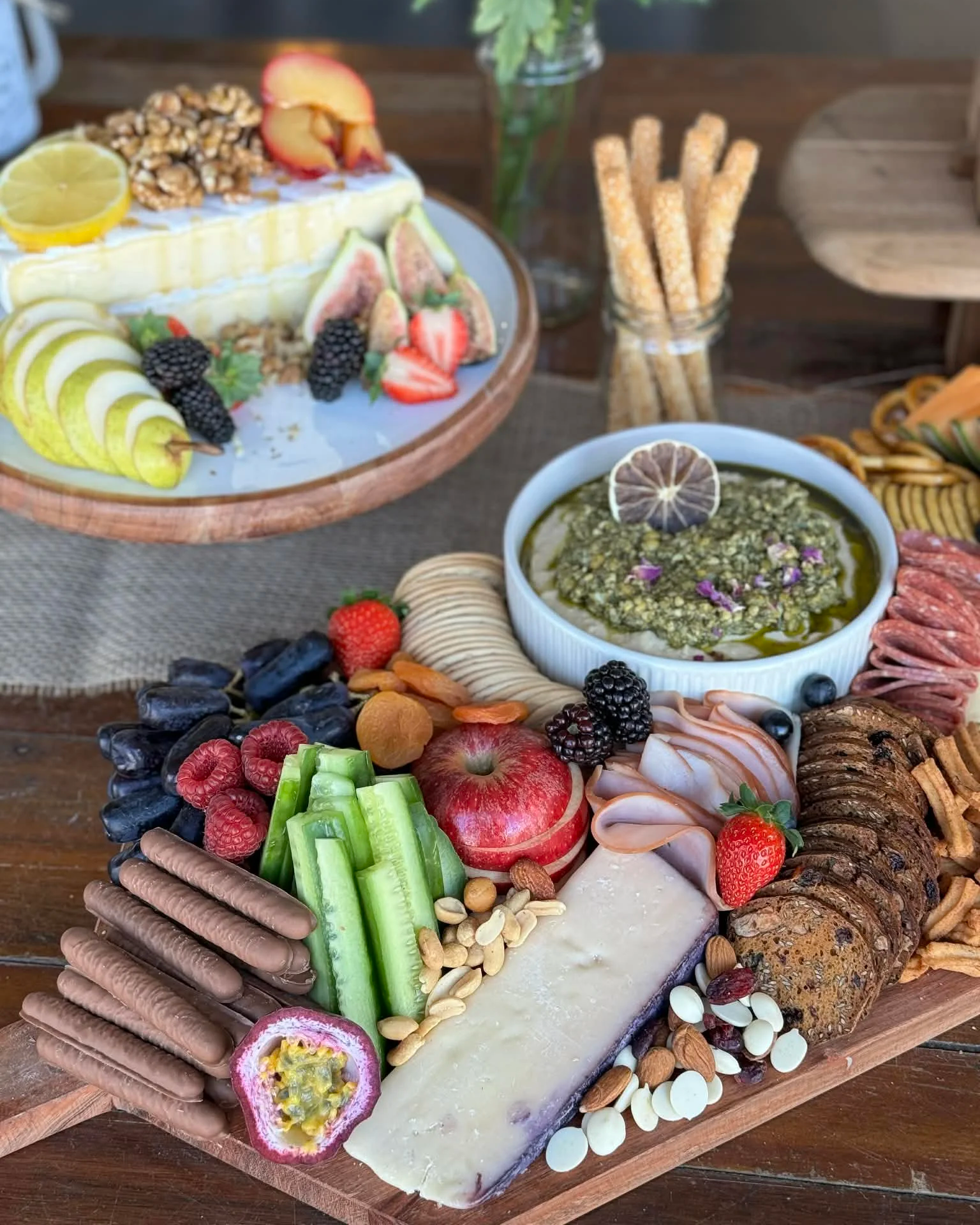 An assortment of snacks including fresh fruits, cheeses, chocolates, nuts, and a bowl of green dip on a wooden platter, arranged for a charcuterie board.