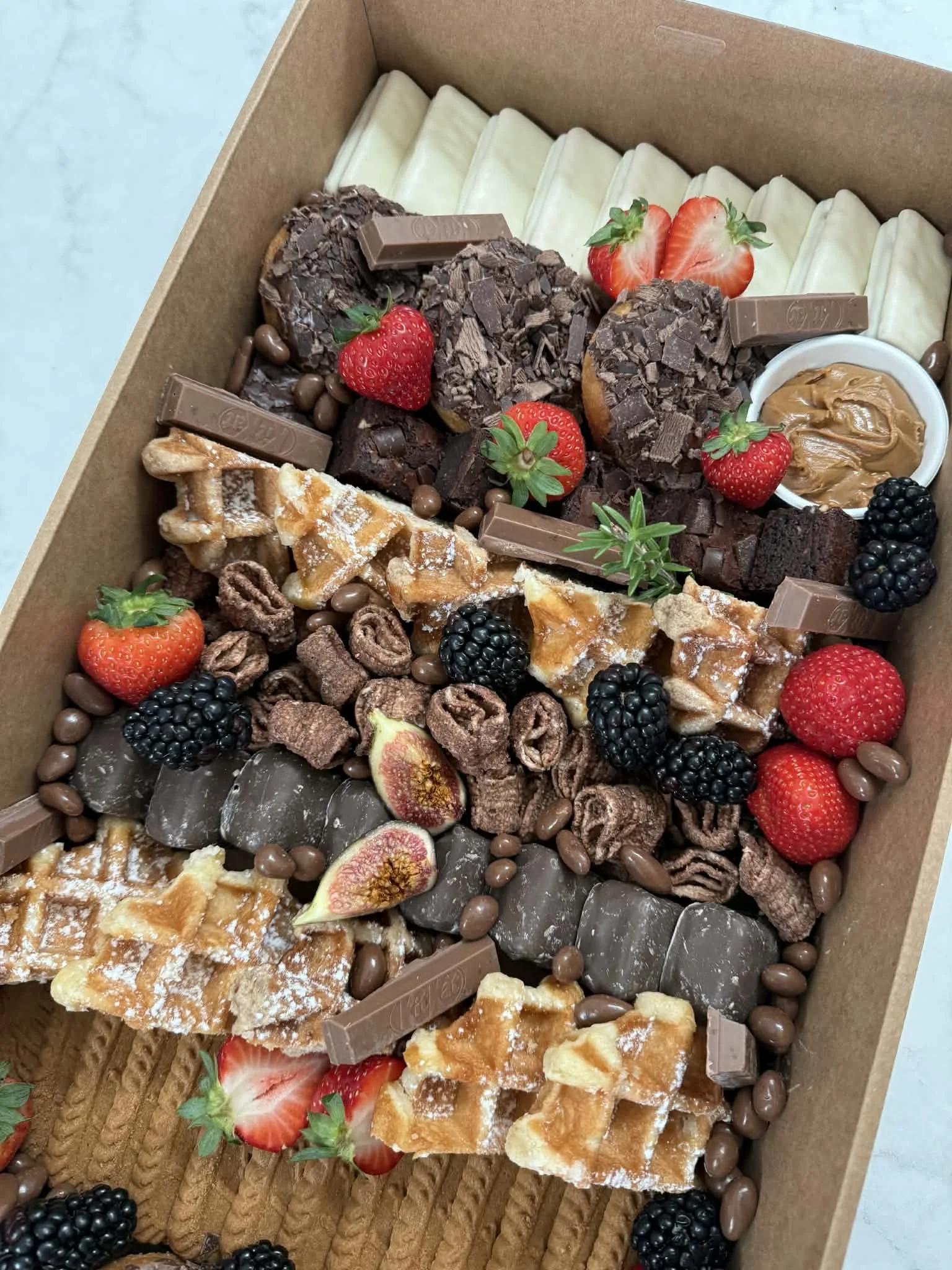 A box of assorted desserts topped with fresh strawberries, blackberries, and figs, including chocolate-covered treats, brownies, cookies, and wafers.