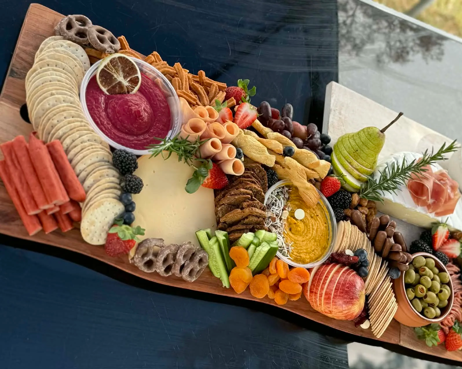 A charcuterie board with assorted cheeses, fruits, crackers, dips, and meats arranged on a wooden serving platter.