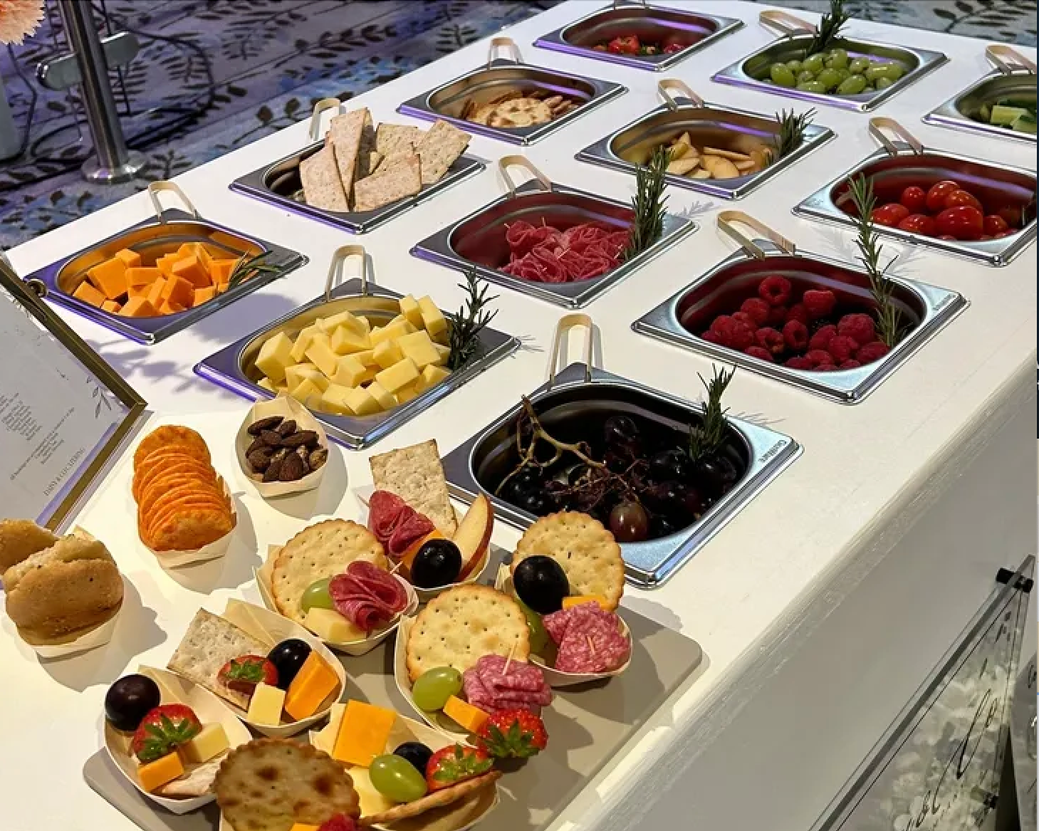 A cheese and fruit platter with crackers, grapes, strawberries, cherry tomatoes, raspberries, and various sliced cheeses. Behind the platter is a salad bar with containers of sliced vegetables, cherry tomatoes, grapes, and cheese, as well as crackers and nuts.