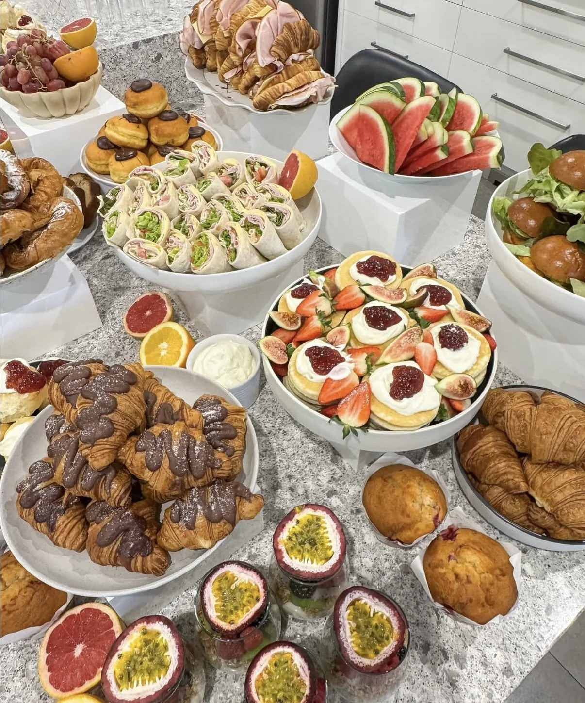 Assorted fruits, pastries, and sandwiches on a kitchen counter including watermelon slices, strawberries with cream, croissants, muffins, and wrap sandwiches.