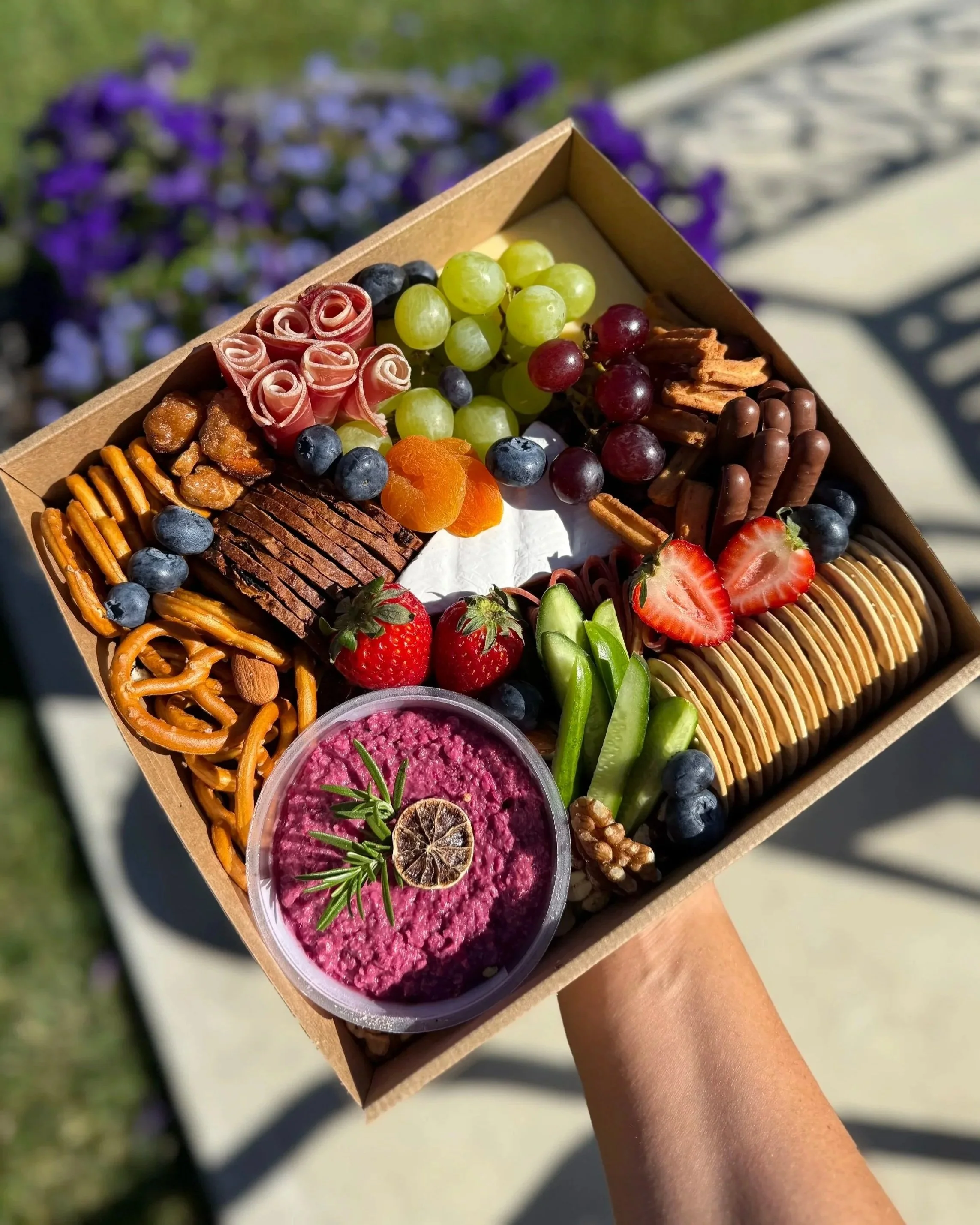 A tray of assorted snacks including grapes, strawberries, sliced strawberries, crackers, pretzels, blue cheese, nuts, dried apricots, chocolate-covered biscuits, and a bowl of pink hummus garnished with a lemon slice and rosemary, held outside in sunlight.