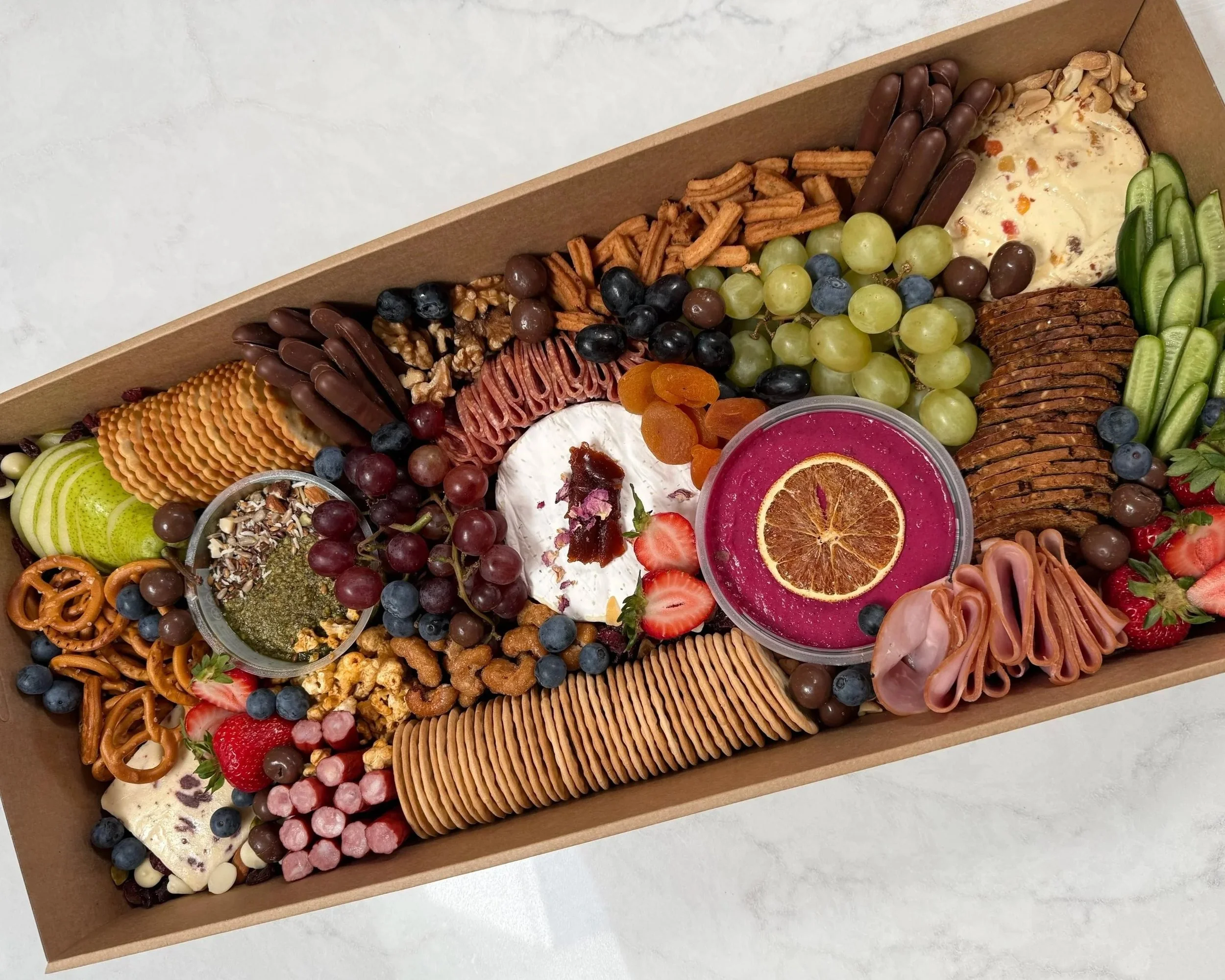 A large assortment of cheeses, meats, fruits, crackers, nuts, and dips in a rectangular box for a charcuterie board.
