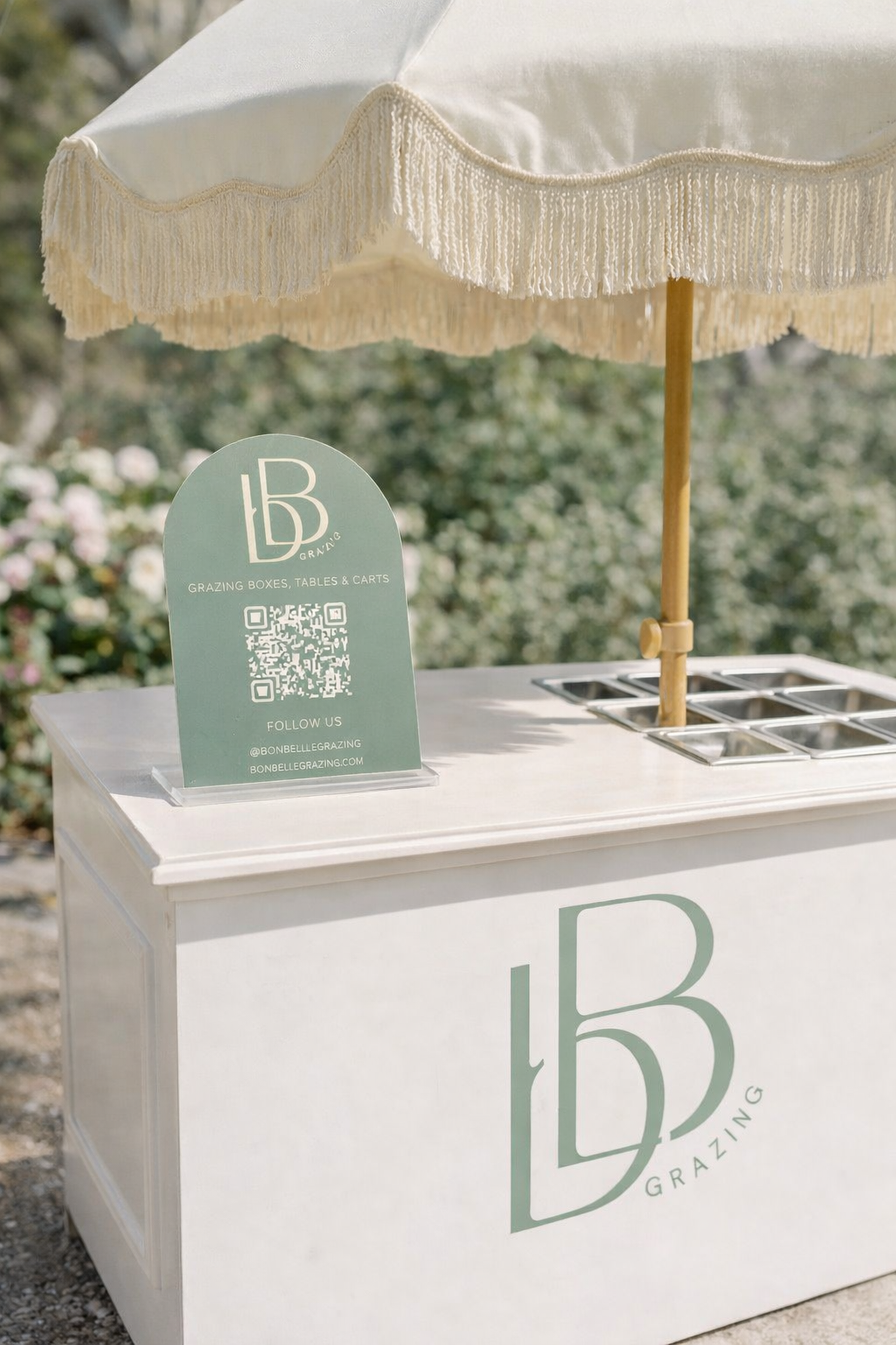 A white outdoor grazing box cart with a large 'LB' Grazing logo and an umbrella, holding small containers for grazing boxes, tables, and carts, with a sign displaying the logo, social media, and a QR code.