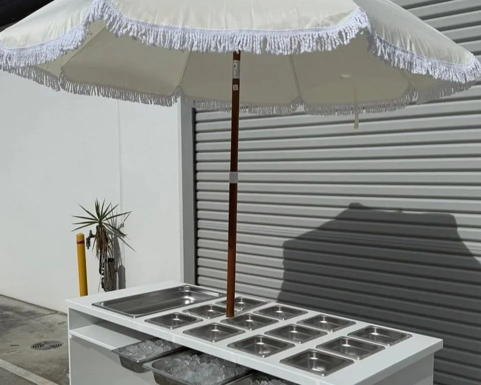 Ice cream or food serving station with multiple stainless steel containers under a large white patio umbrella.