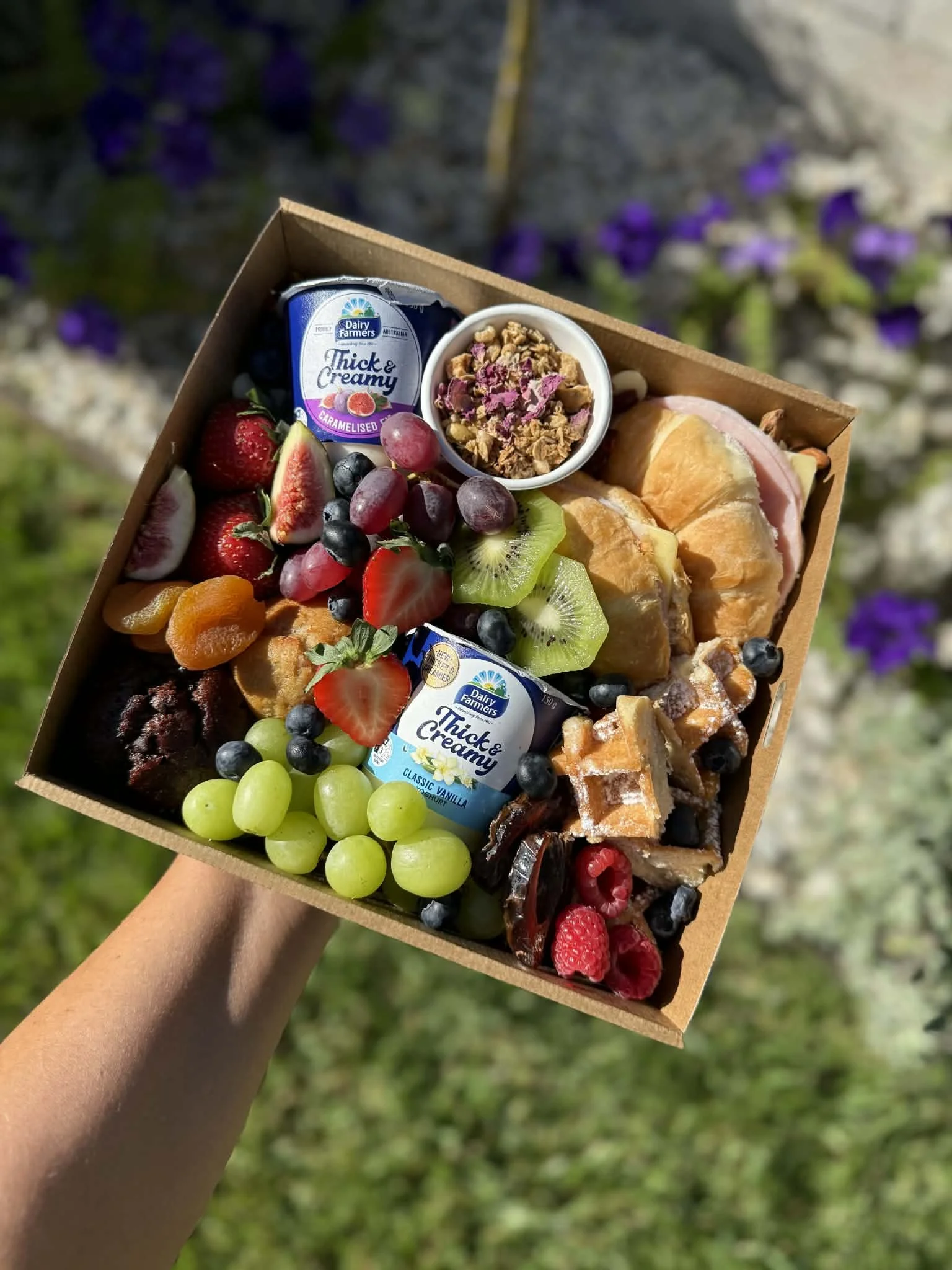 A hand holding a box filled with a variety of fresh fruit, granola, and two small containers of dessert, set outdoors with purple flowers in the background.