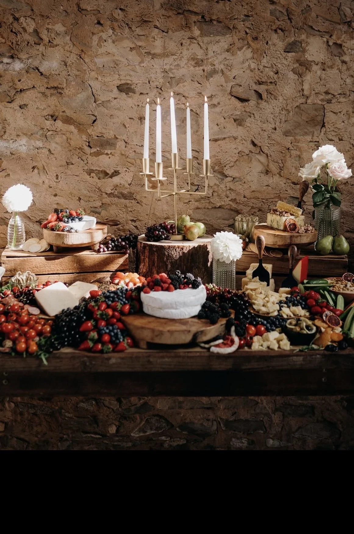 A rustic table with cheese, berries, fruit, flowers, and a modern candle holder against a stone wall.