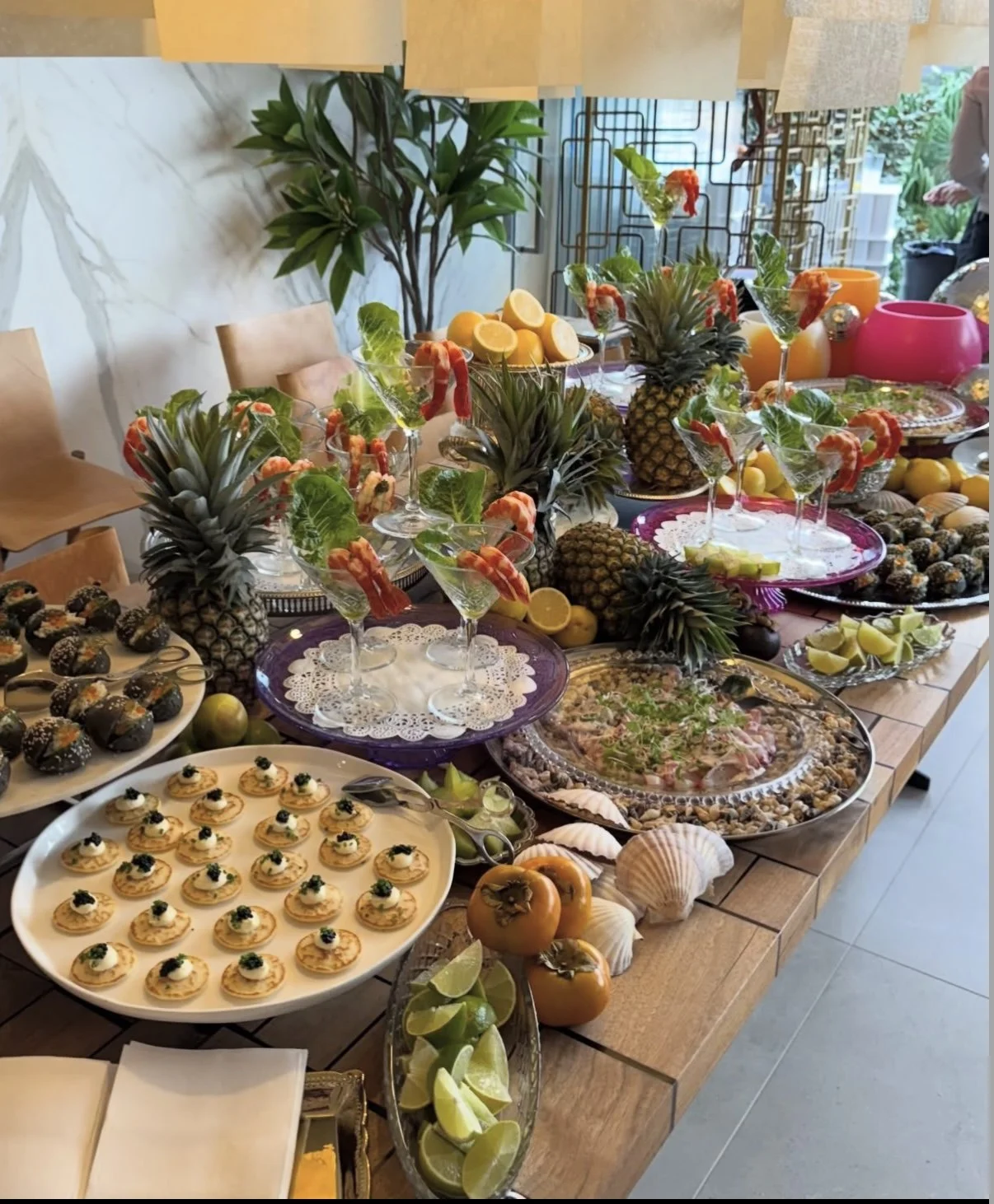 A table decorated with fresh pineapples, lemons, and tropical fruits with various appetizers and snacks including mini canapés, seafood, and desserts arranged for a celebration or party.