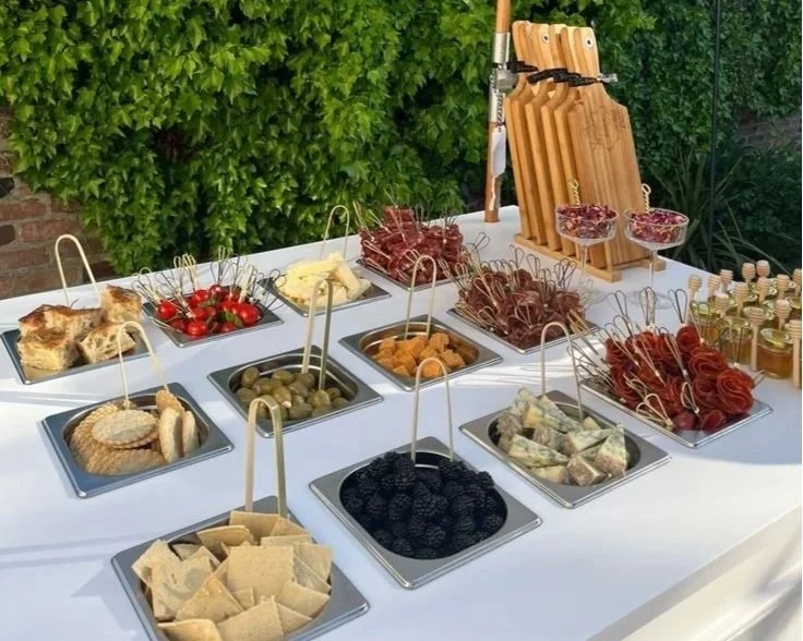 Assorted appetizers and snacks on a white table, including cherry tomatoes, crackers, cheese, olives, breaded items, dried meat, and assorted small jars, with wooden cutting boards and knives in the background