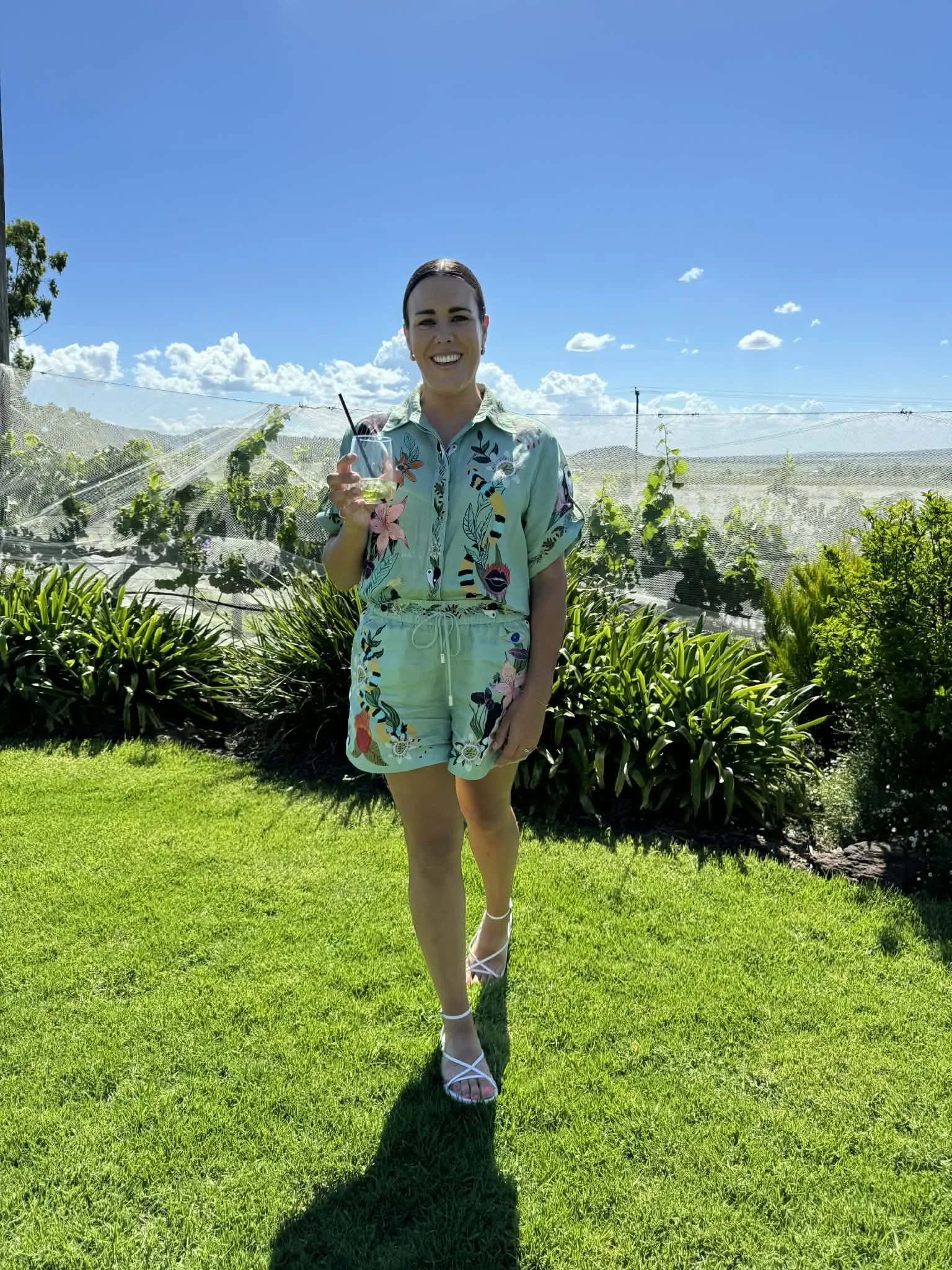 A woman in summery floral outfit holding a drink, standing on a lush green lawn with greenery and blue sky in the background.