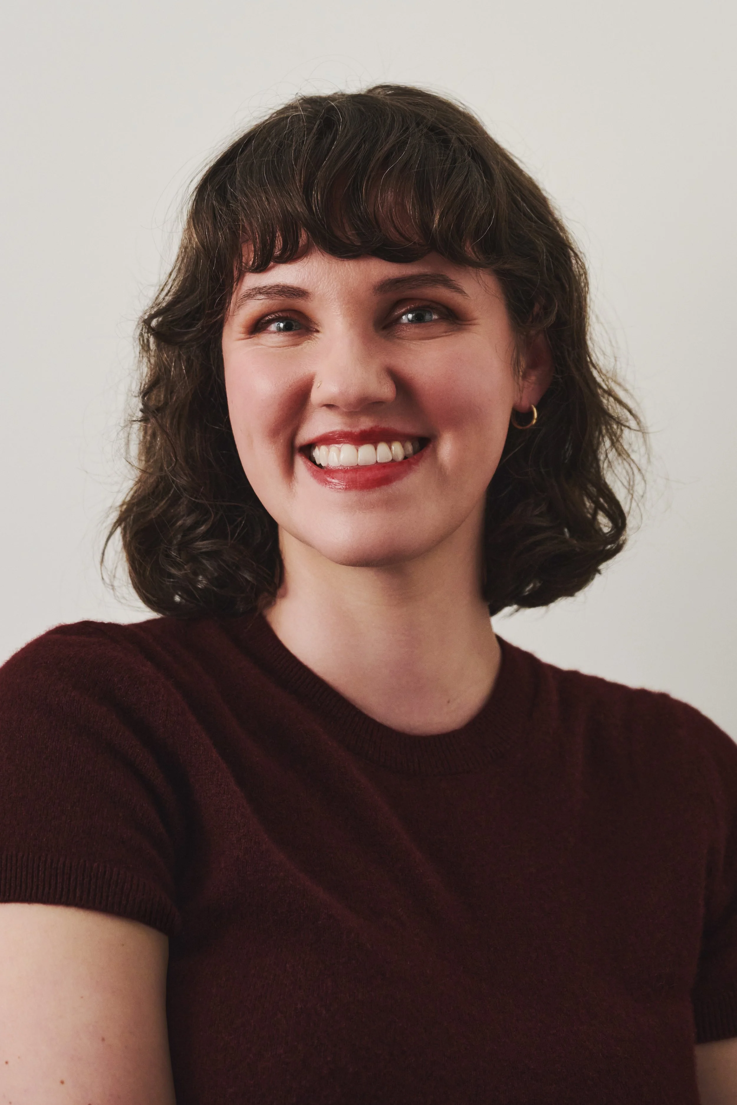 Close-up of a young woman with wavy brown hair, wearing a maroon shirt, smiling against a plain background.