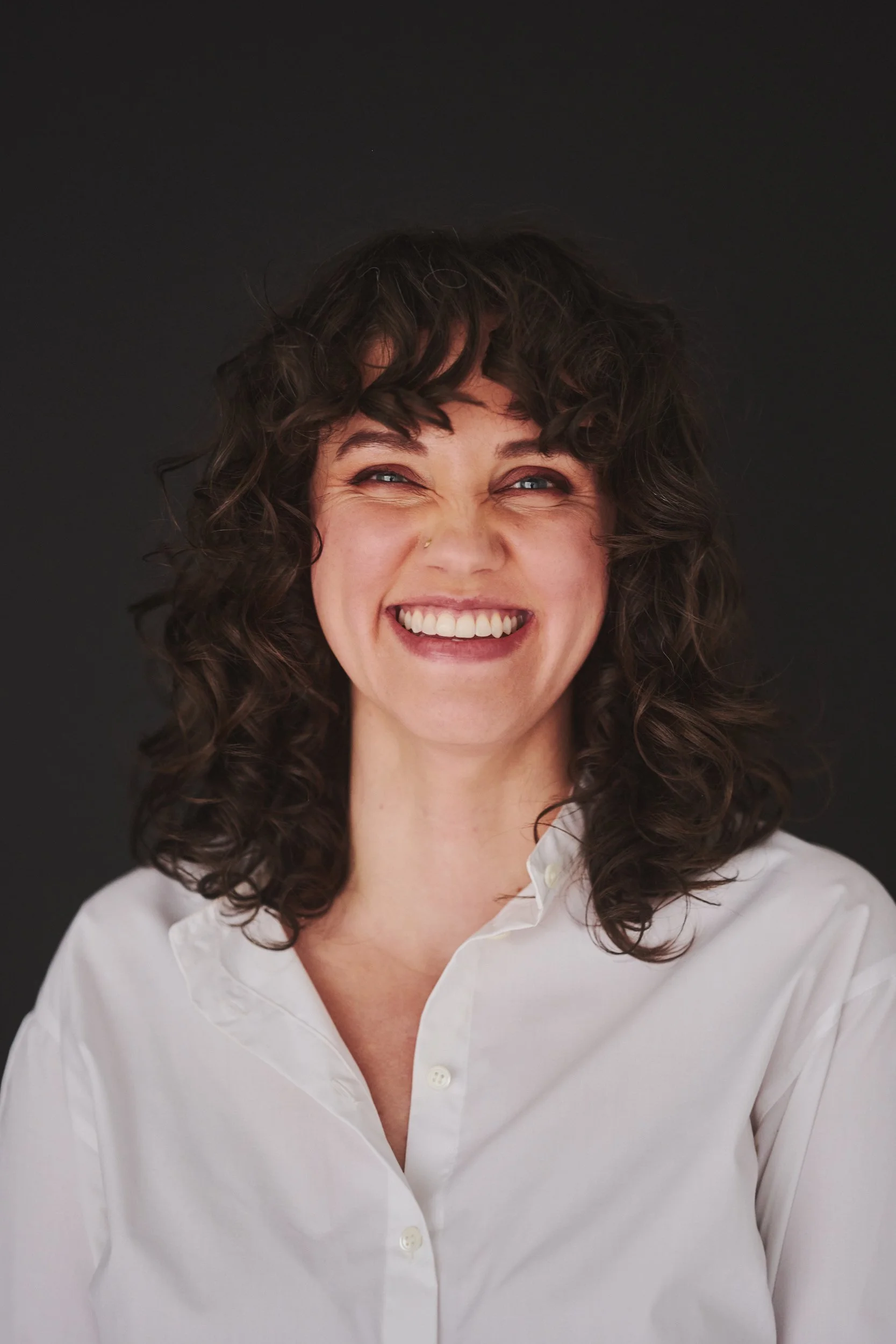 A woman with curly dark hair smiling broadly in front of a black background.