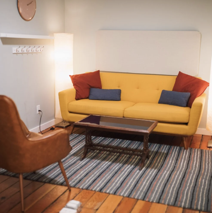 Living room with a yellow couch, red and blue pillows, a wooden coffee table, a tan armchair, a striped rug, a floor lamp, and a gray wall with a clock and coat hooks.