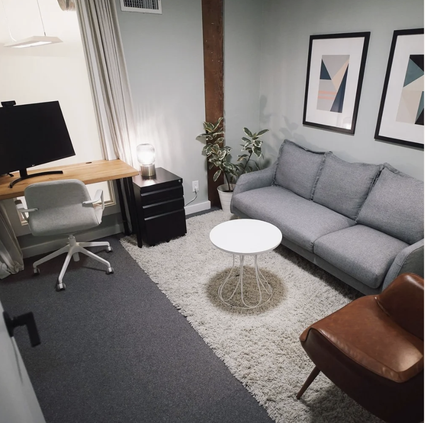 A cozy room with a gray sofa, a white round coffee table, and a brown leather armchair. There's a potted plant beside the sofa, framed geometric art on the wall, a small black side table, a lamp, a desk with a monitor, and a white office chair.