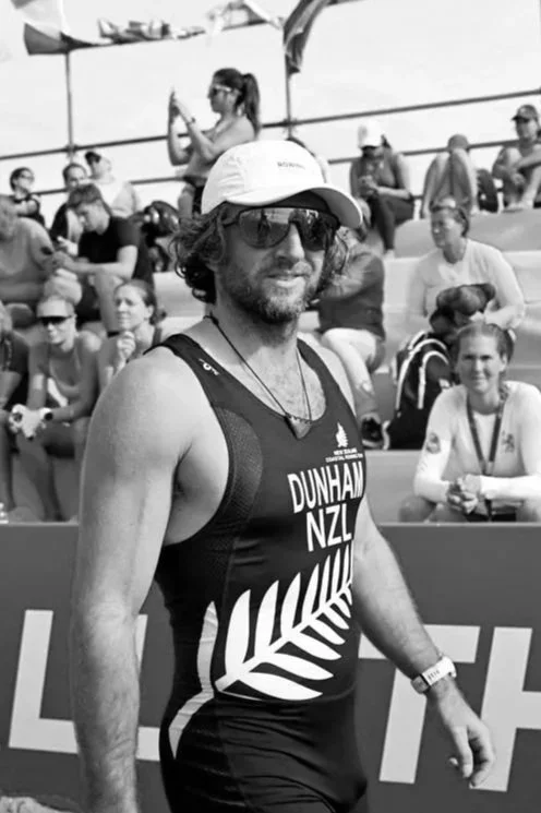 Male athlete at a track and field event, wearing sunglasses, a hat, and a sports uniform with 'Dunham NZL' printed on it, standing in front of a crowd of spectators.