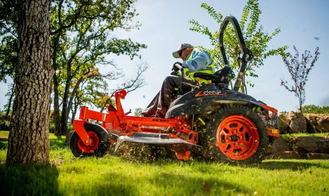 A person operating a riding lawn mower on a grassy area, surrounded by trees and rocks, with a clear blue sky.