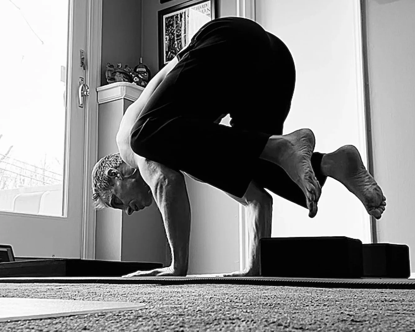 A person practicing yoga indoors, balancing on one hand with their legs bent and head down, near large windows and indoor decor.
