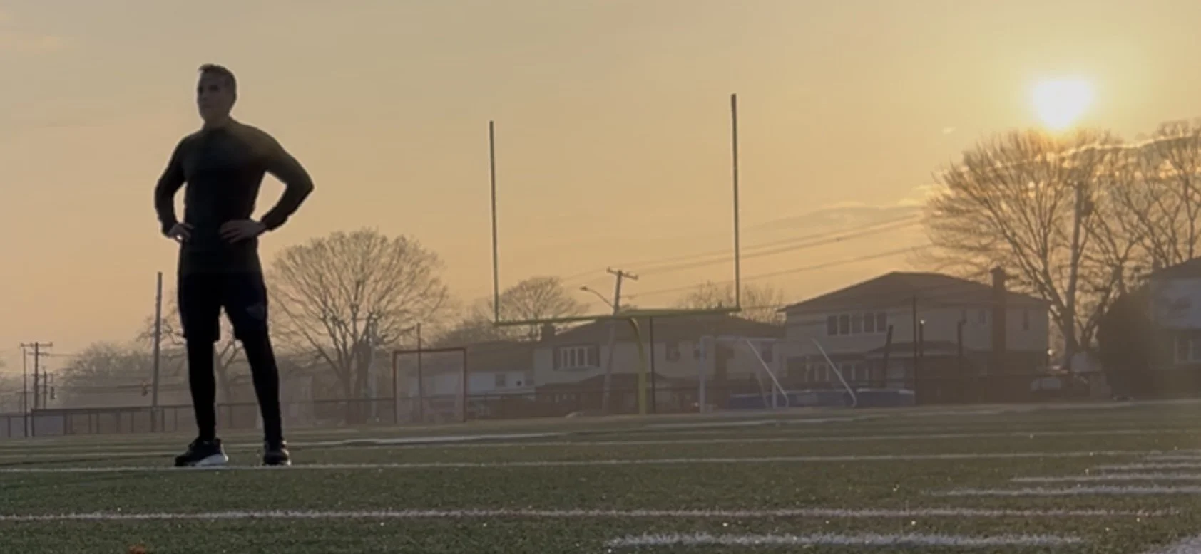 A person standing on a football field at sunset, wearing black athletic clothing, with trees and houses in the background.