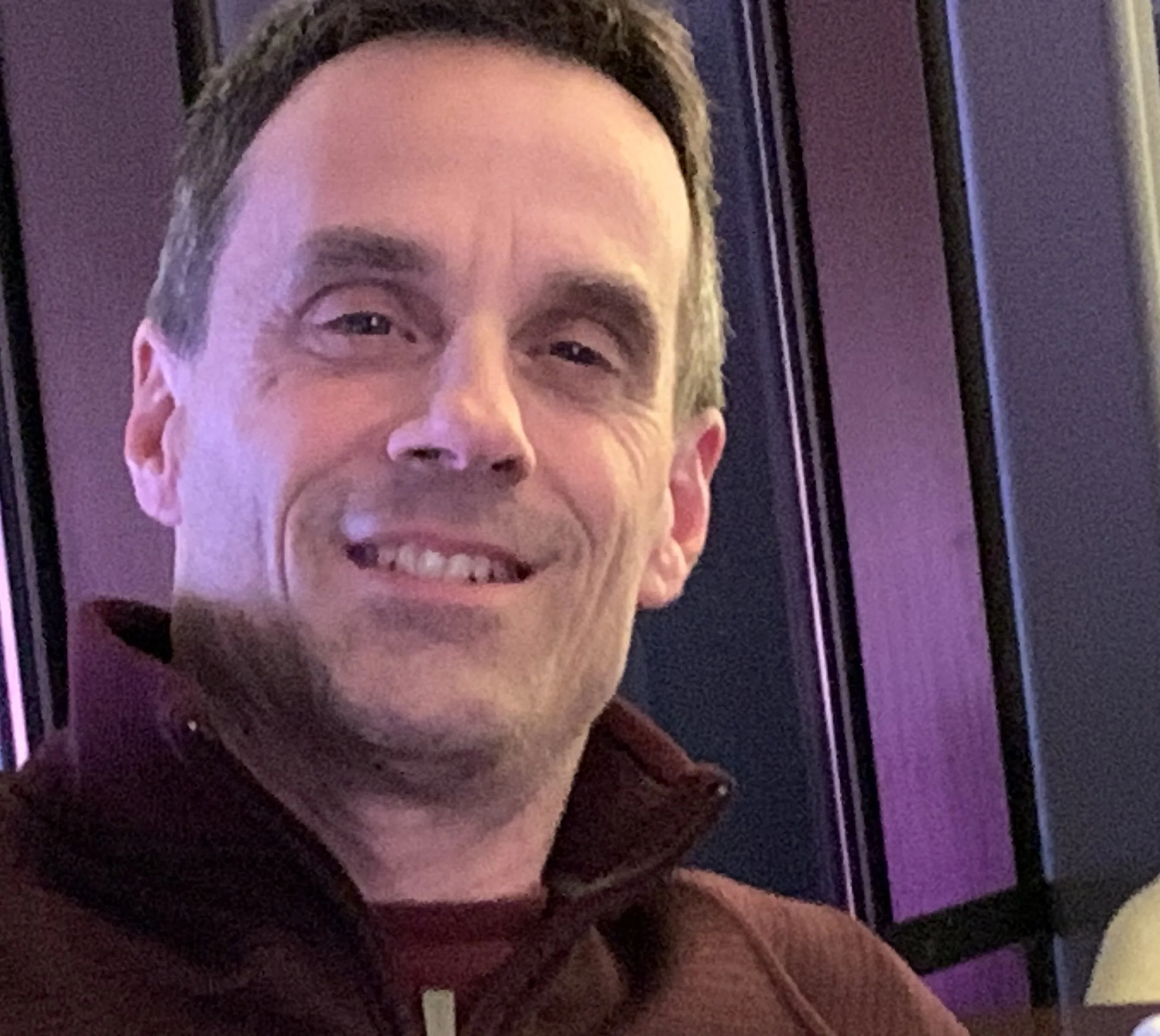 Close-up of a smiling man wearing a black beanie and a maroon hoodie, with blurred purple and yellow background.