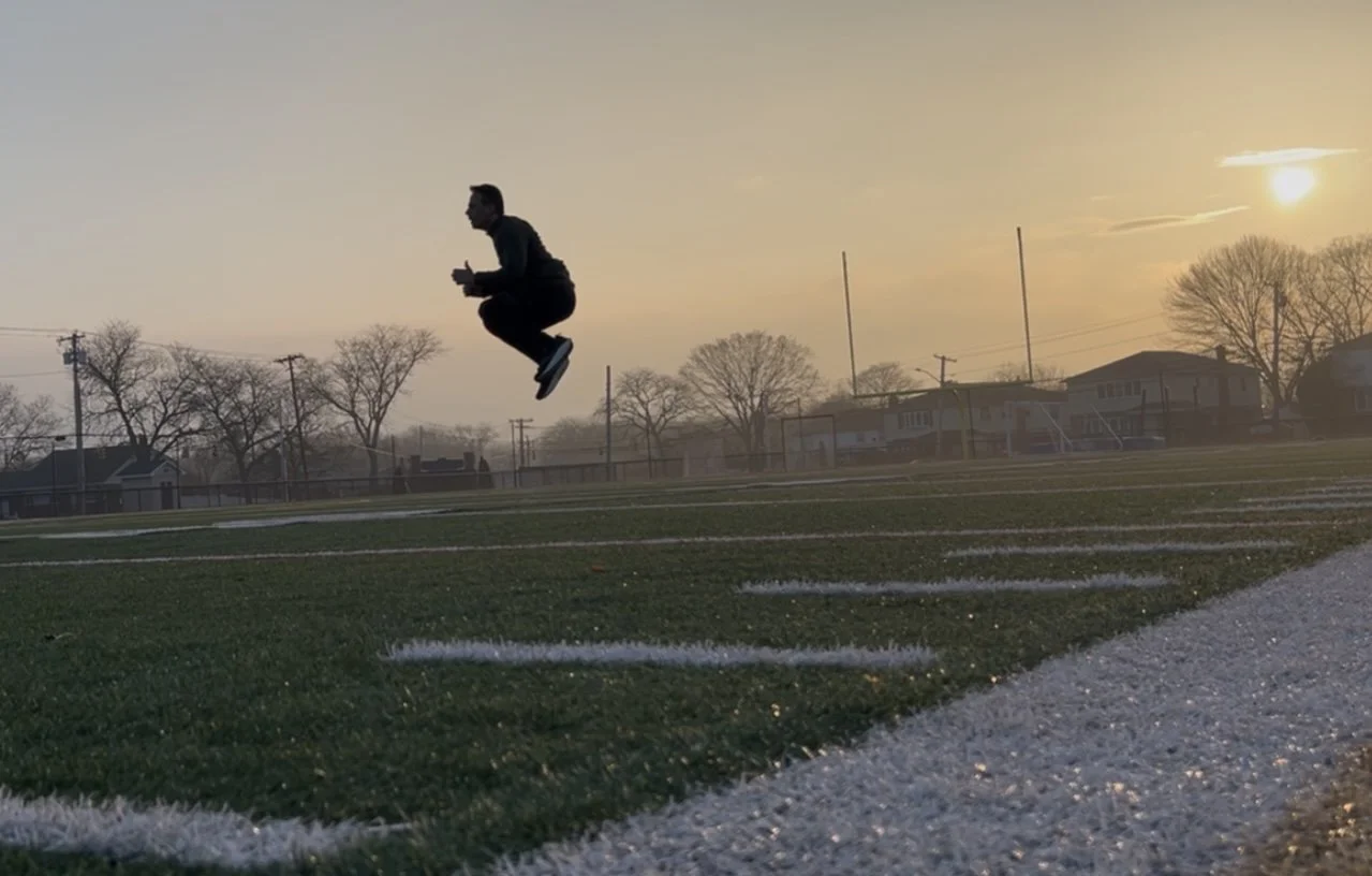 Person jumping in the air on a soccer field during sunset with empty goalposts and houses in the background.