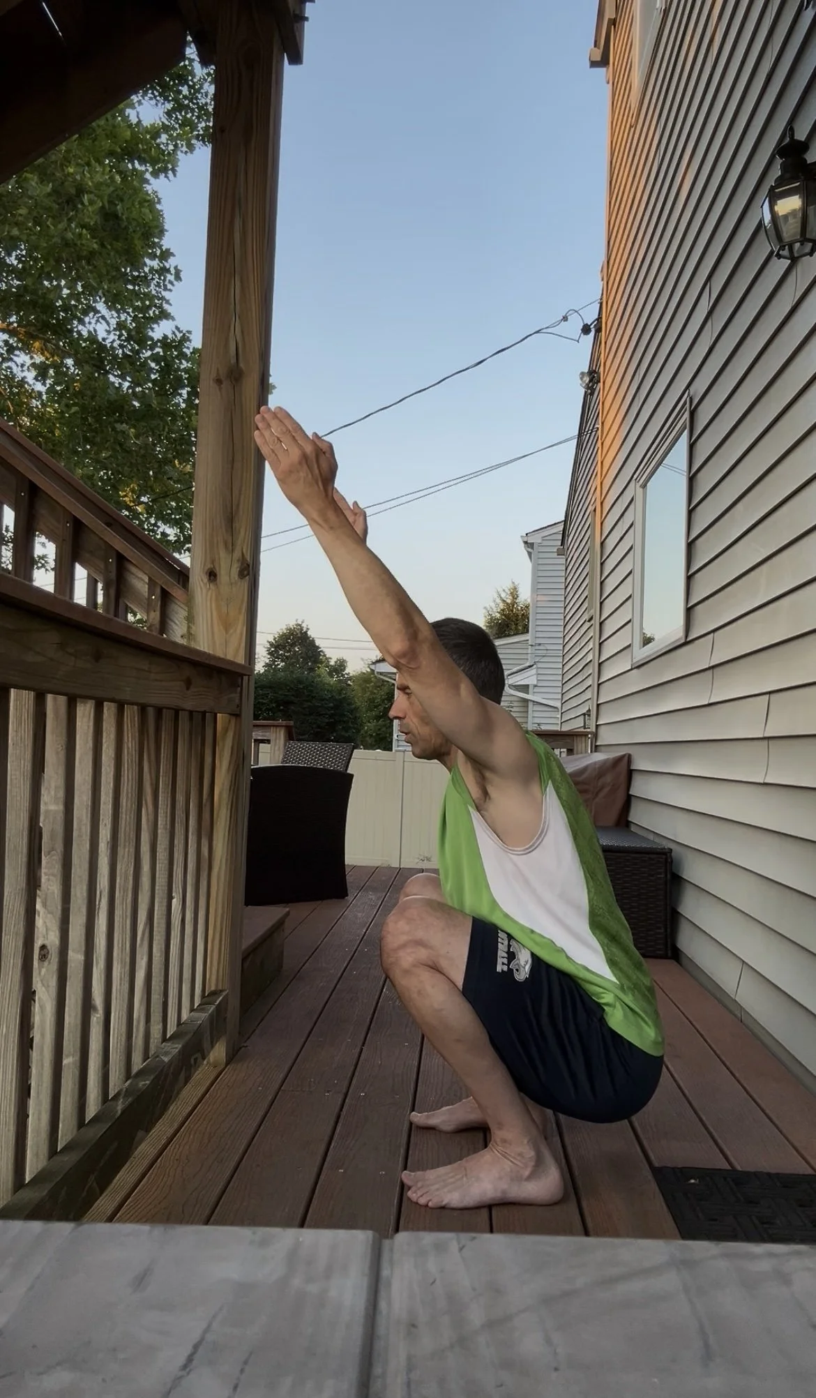 A man in a green and white sleeveless shirt and black shorts is performing a squat on a wooden deck outside a house. He has his hands raised above his head and is looking downwards.