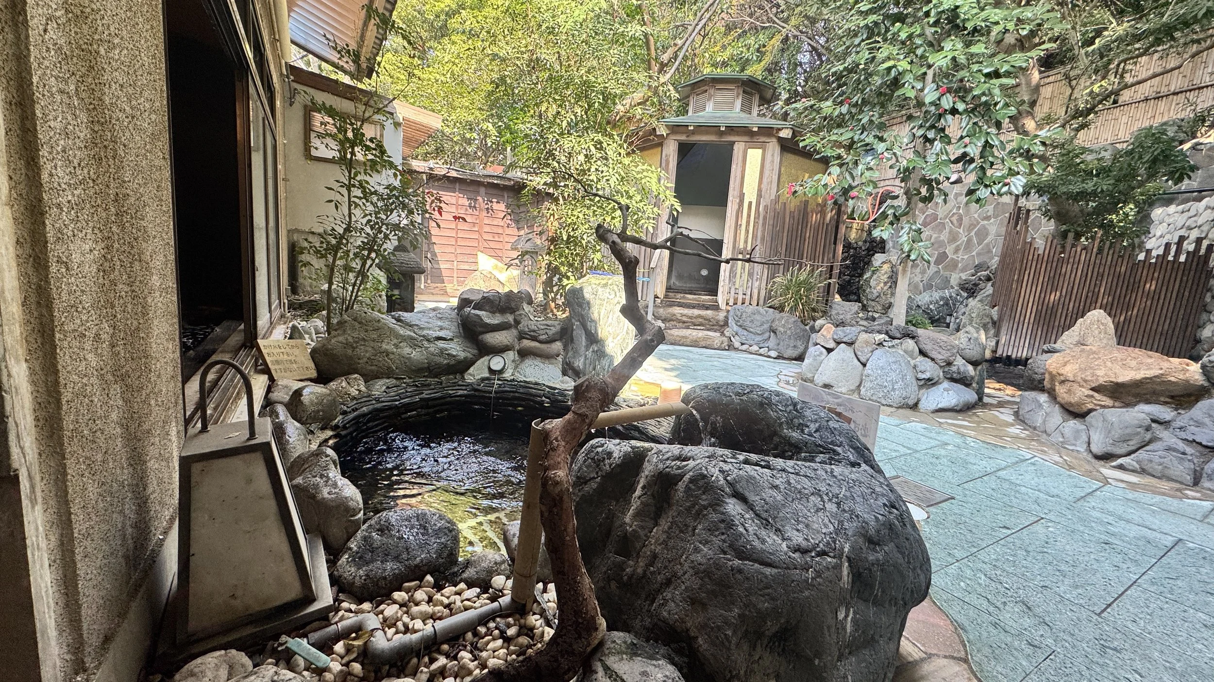 屋外の温泉の景色。岩や石、木々に囲まれた静かな空間。温泉の水面が見える。