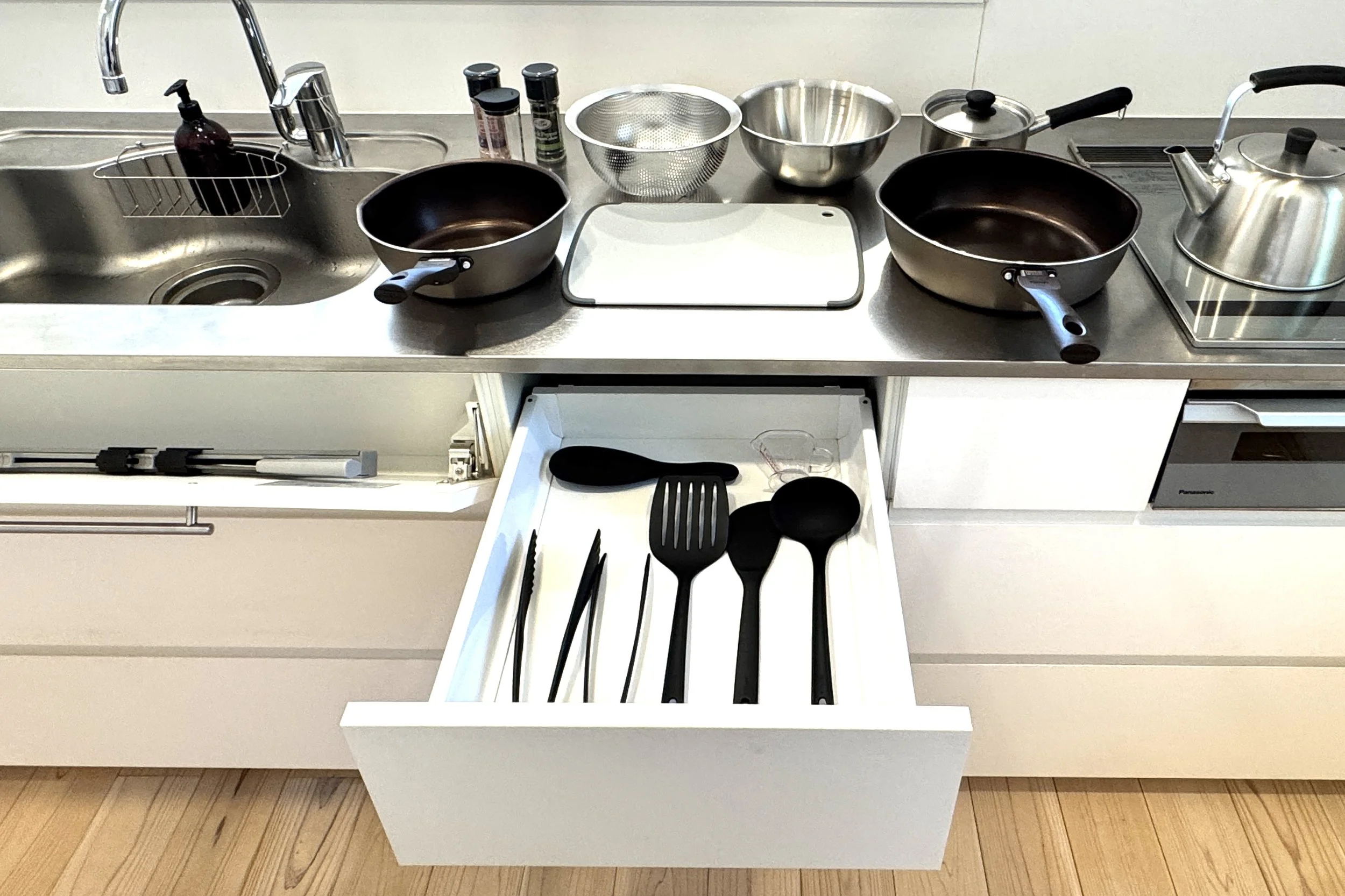 引き出しの中にキッチングッズと調理器具が収納されているアイランドキッチンの写真。シンクには洗剤とスポンジ、ステンレスのザルや鍋、計量カップなどが配置されている。