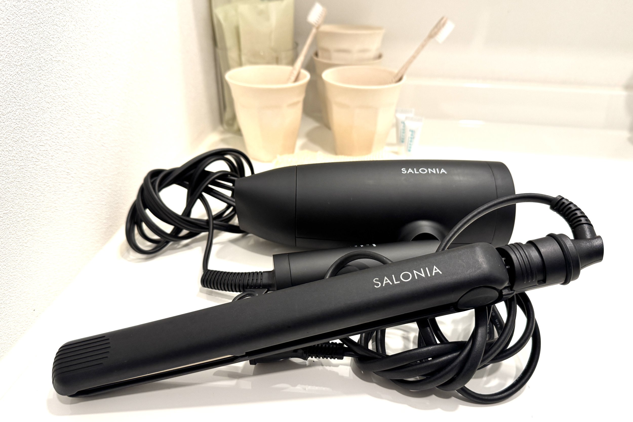 美容師用のヘアアイロンと電源ケーブル、背景に紙コップと歯ブラシ、壁に突っ張り棒が見える画像