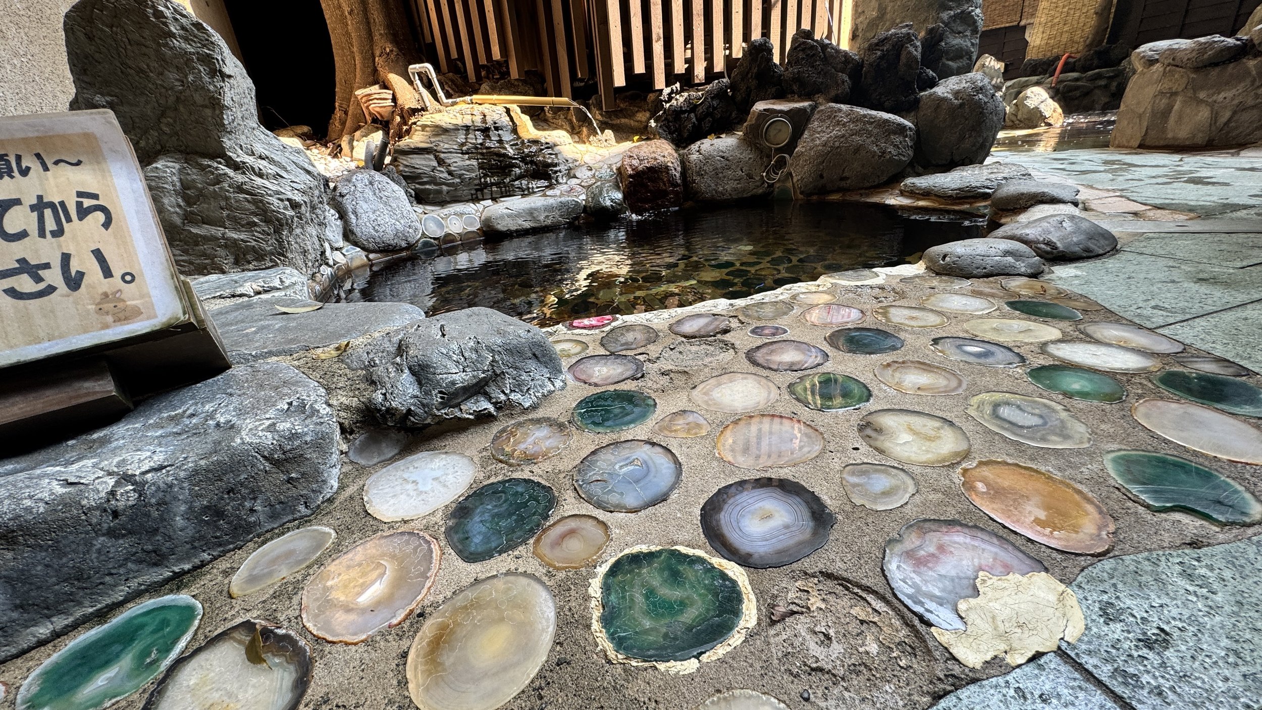 露天風呂の岩とタイルの浴場。中央に黒い温泉や水の湯が張られている。壁や床にはさまざまな色の貝殻が埋め込まれている。