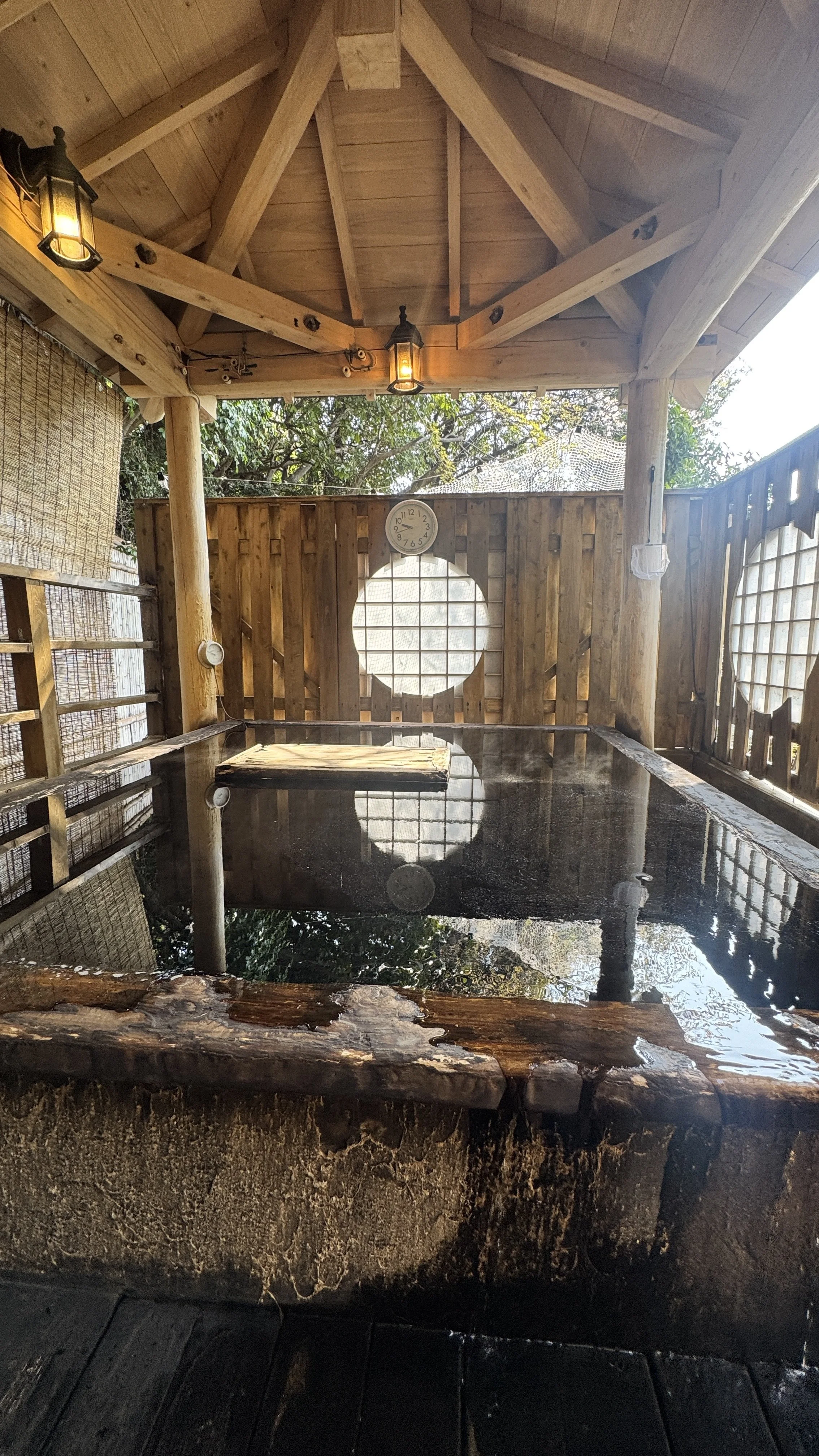 木製の屋根と壁に囲まれた露天風呂。壁には時計が掛かっており、自然の風景とともに暖かい照明が設置されている。