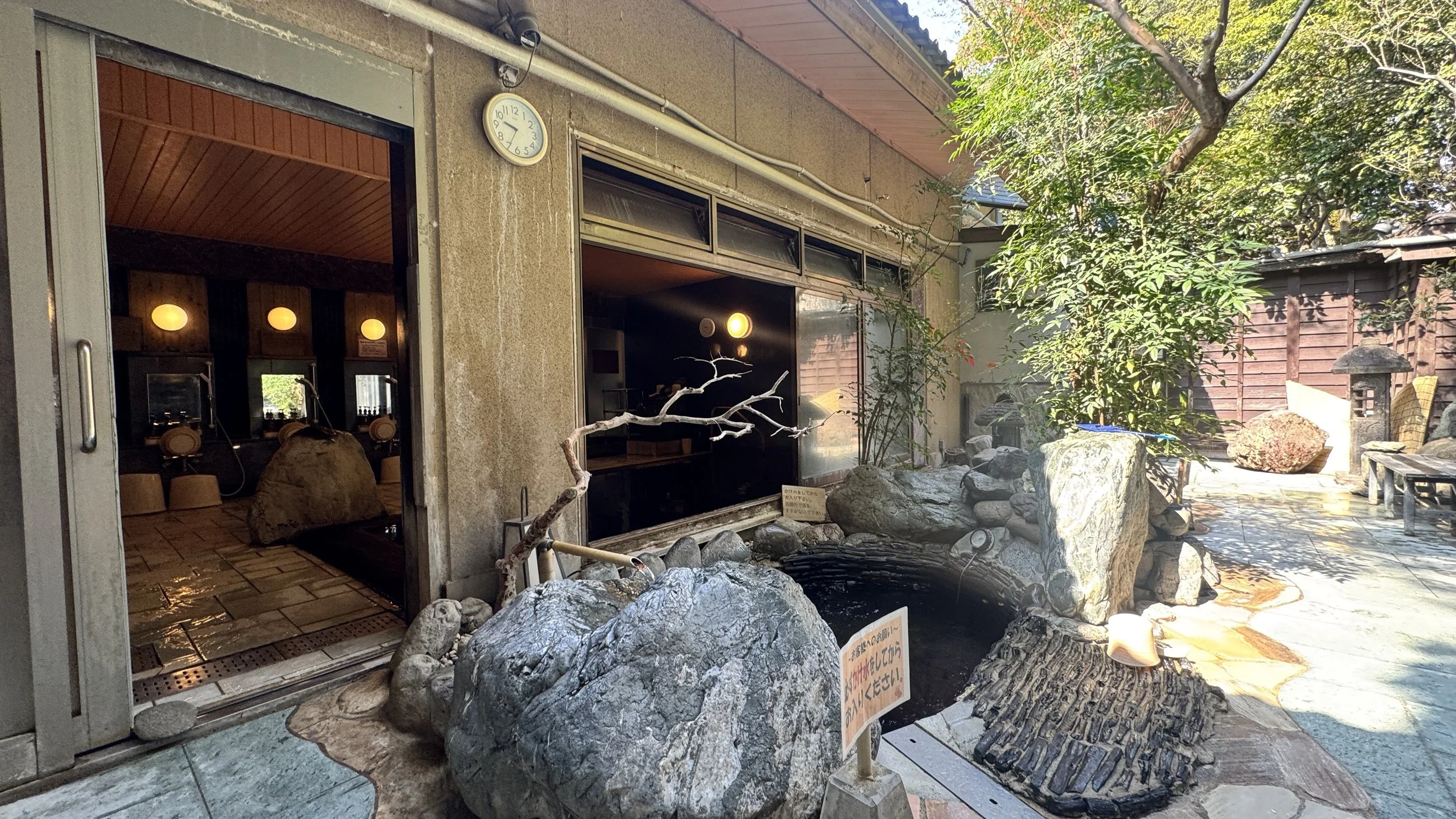 岩や木々に囲まれた日本庭園の人工の池と岩風呂。木の葉が茂る庭園、木造の建物の一部が見える。