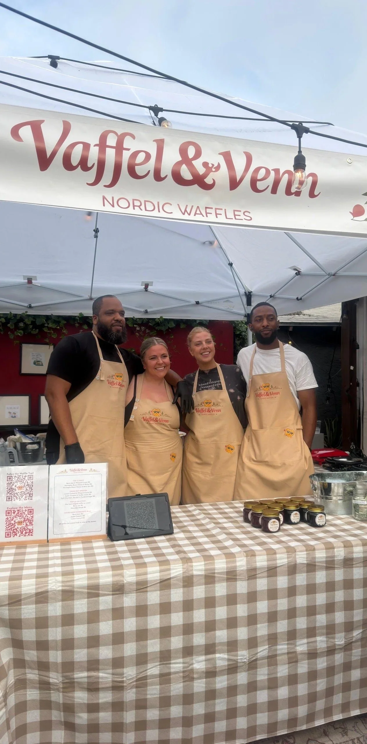 Four people standing behind a table at a Nordic waffle stand, all wearing aprons with the Vaffel & Venn logo, smiling, in front of a red wall and a white canopy.
