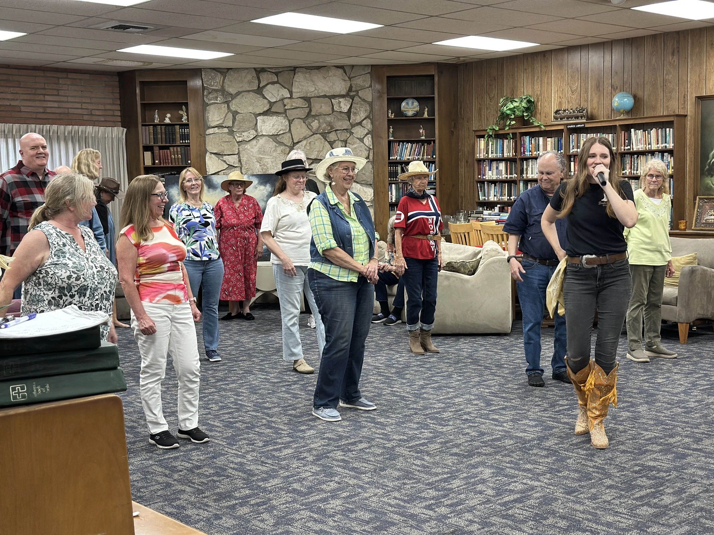 Group of adults gathered indoors for a line dancing class, led by a young woman holding a microphone, wearing a black shirt and cowboy boots.