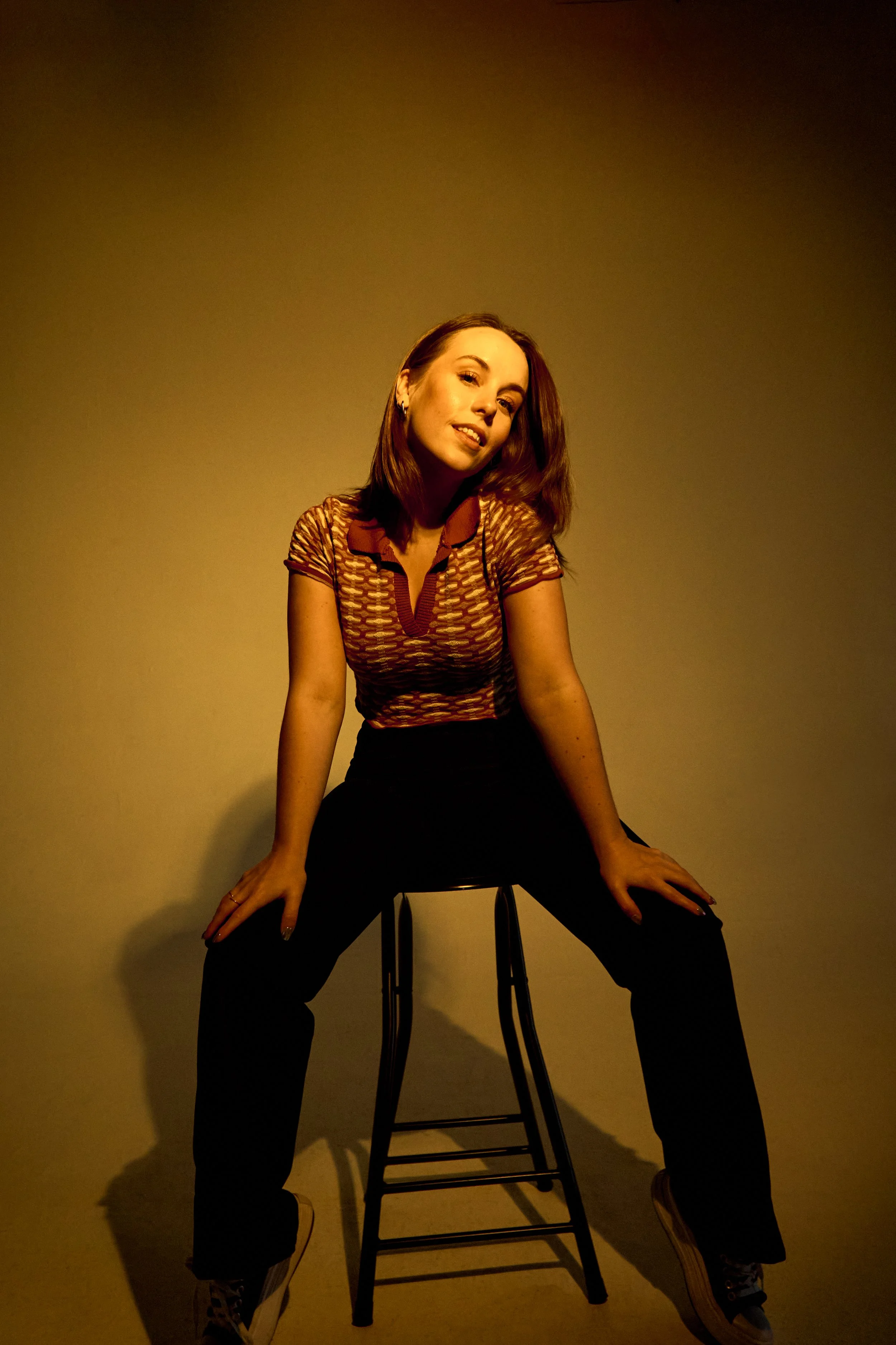 A woman with shoulder-length brown hair, wearing a patterned short-sleeve shirt and black pants, sitting on a stool with her legs apart and hands resting on her thighs against a plain beige wall with lighting casting shadows.